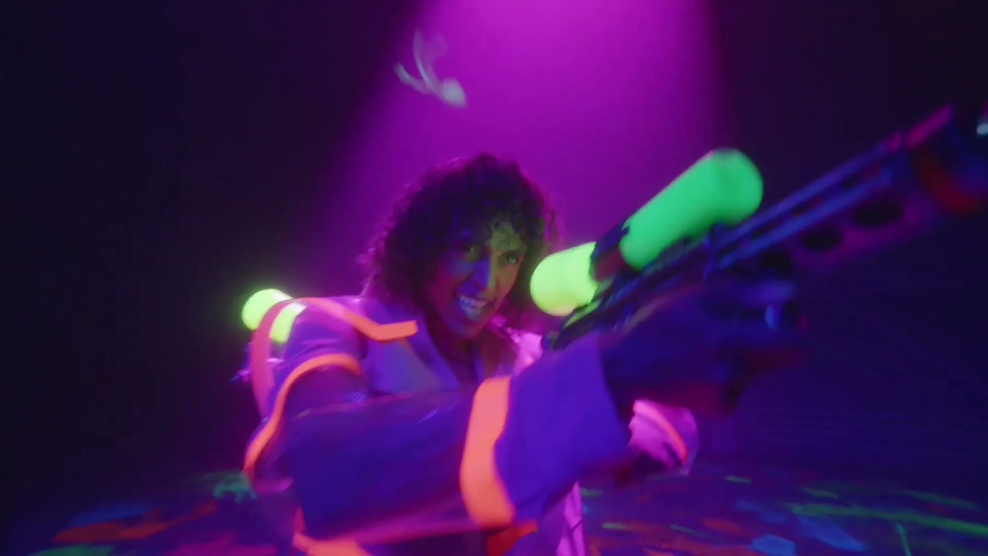 A young woman with curly hair, wearing a futuristic neon outfit, aims a glowing laser gun in a dark, neon-lit room, as if in a video game.