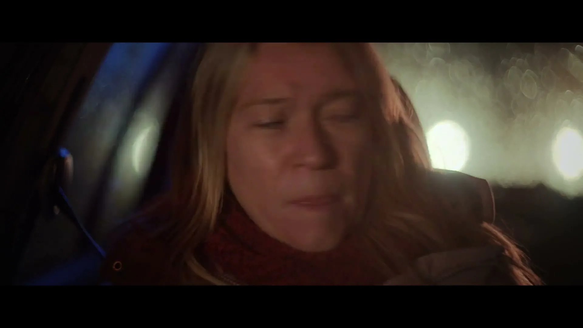 A blonde woman with a red scarf looks distressed while sitting in a car at night, with blurry lights visible through the window.