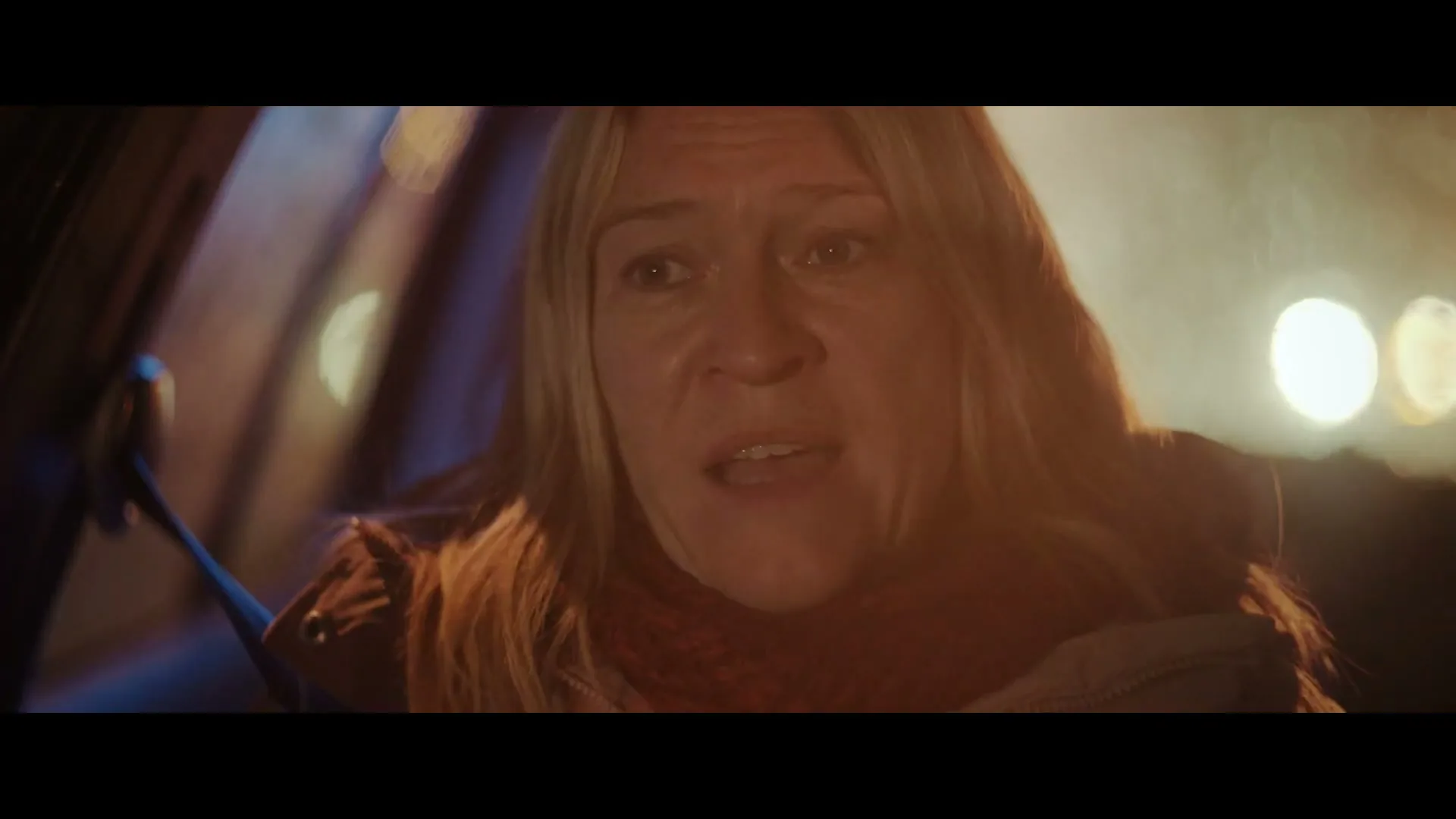 A woman with blonde hair and a red scarf looks distressed while sitting in a car at night, with blurry lights in the background.