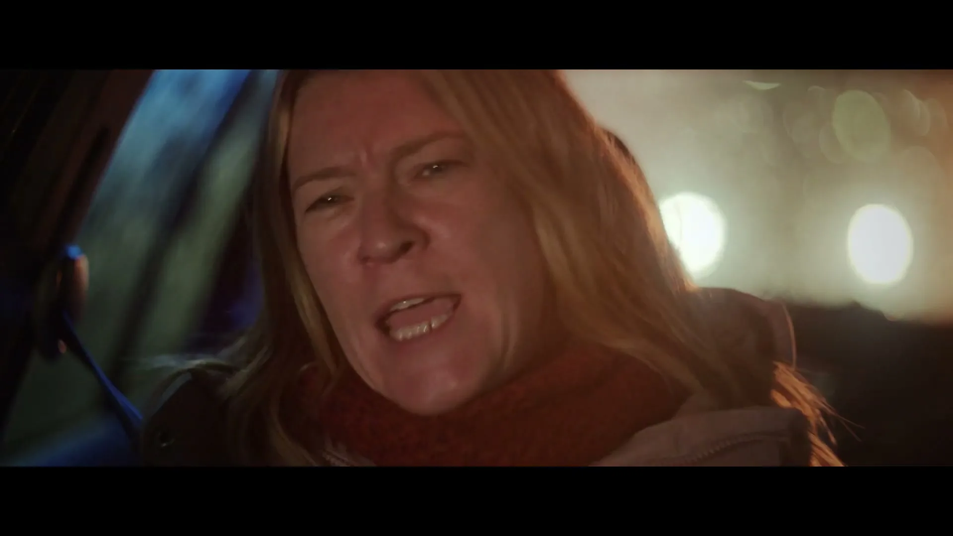 A blonde woman in her 40s, wearing a red scarf and winter coat, screams in frustration while driving at night.
