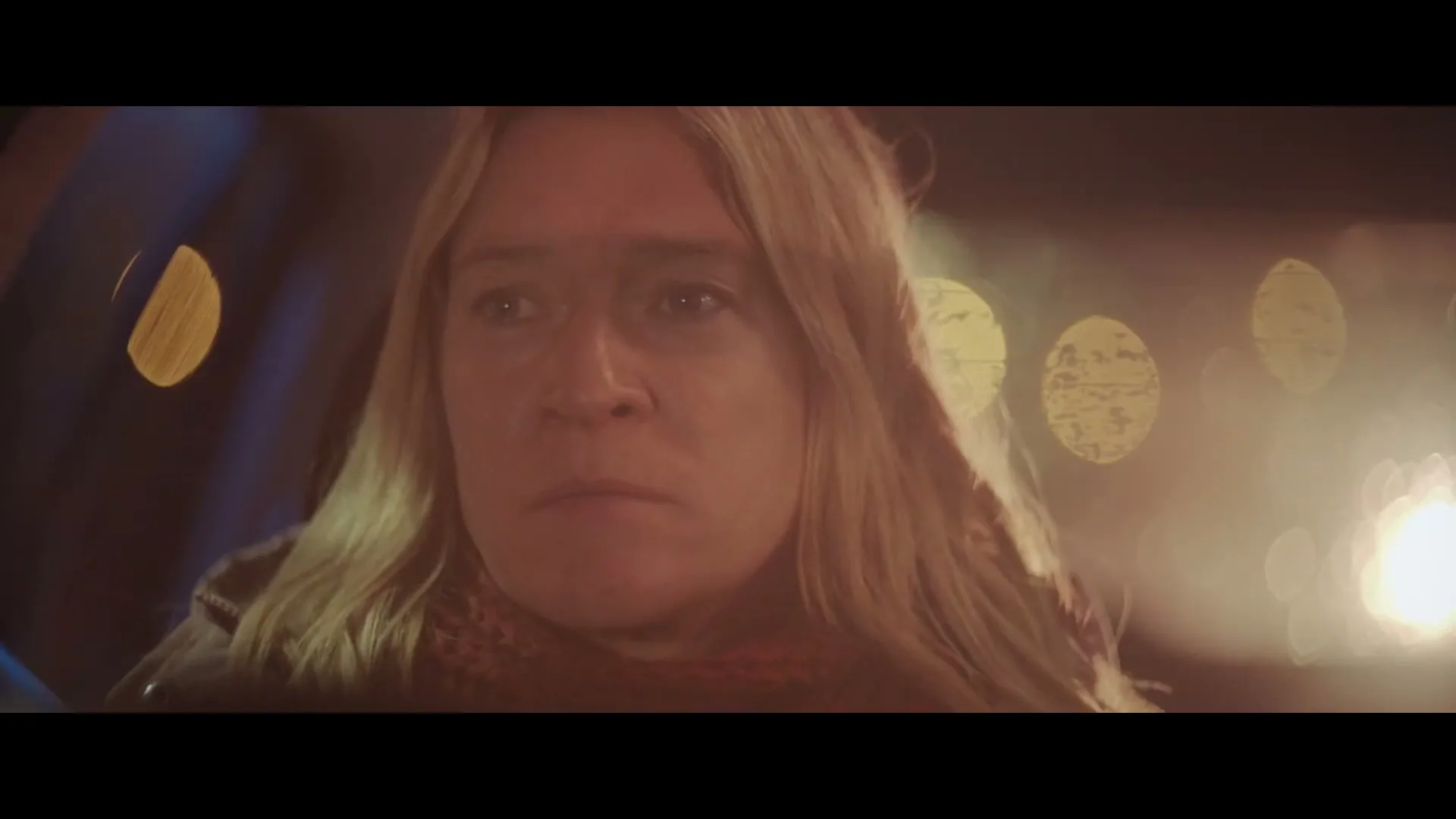 A woman with blonde hair and a red scarf looks worried while sitting in a car at night, with bokeh lights in the background.