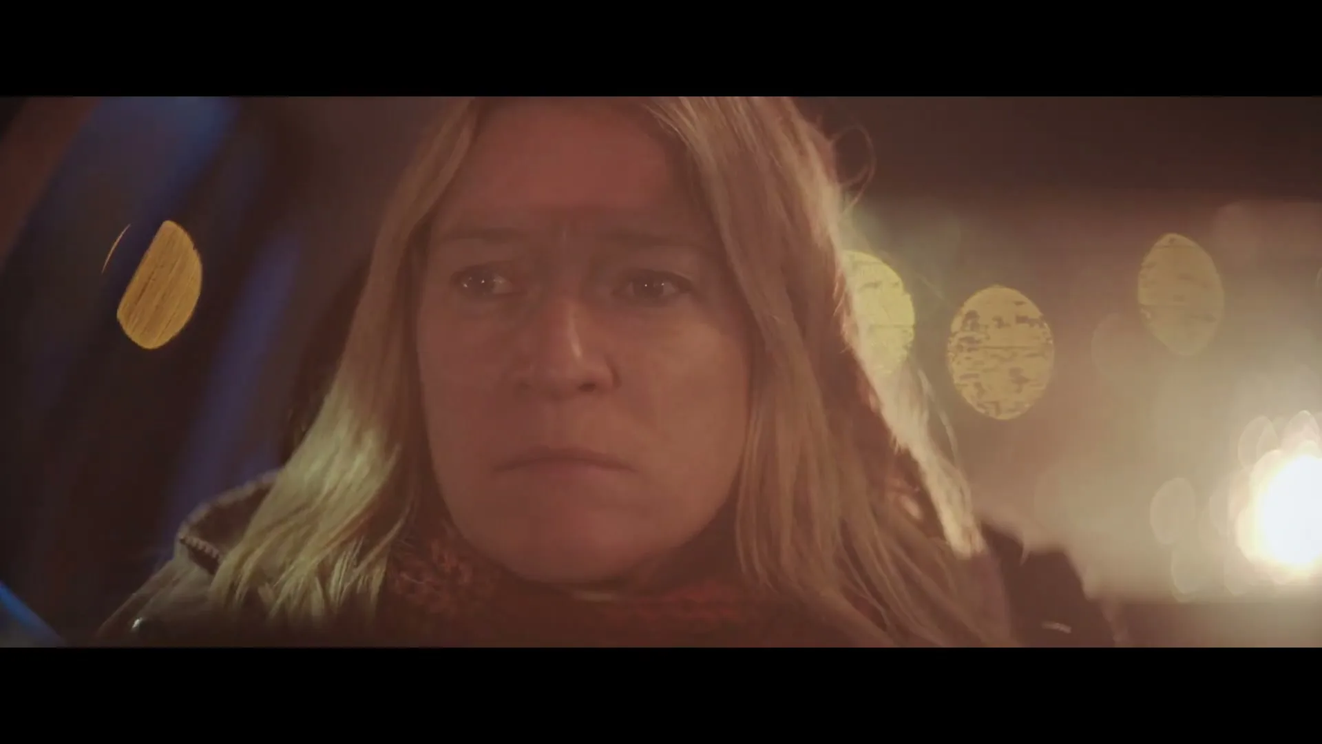 A blonde woman with a worried expression sits in a car at night, with blurry lights visible through the window.