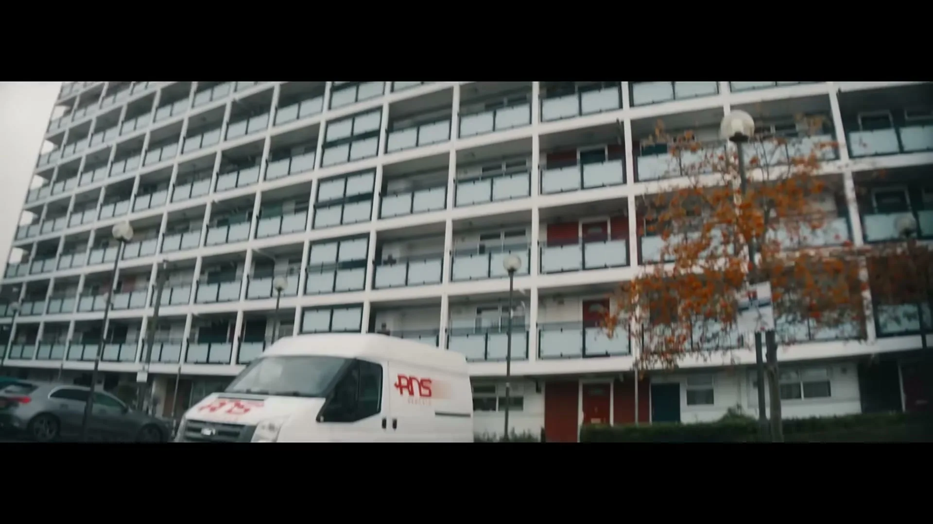 A white delivery van with red lettering drives past a large, multi-story apartment building on an overcast day.