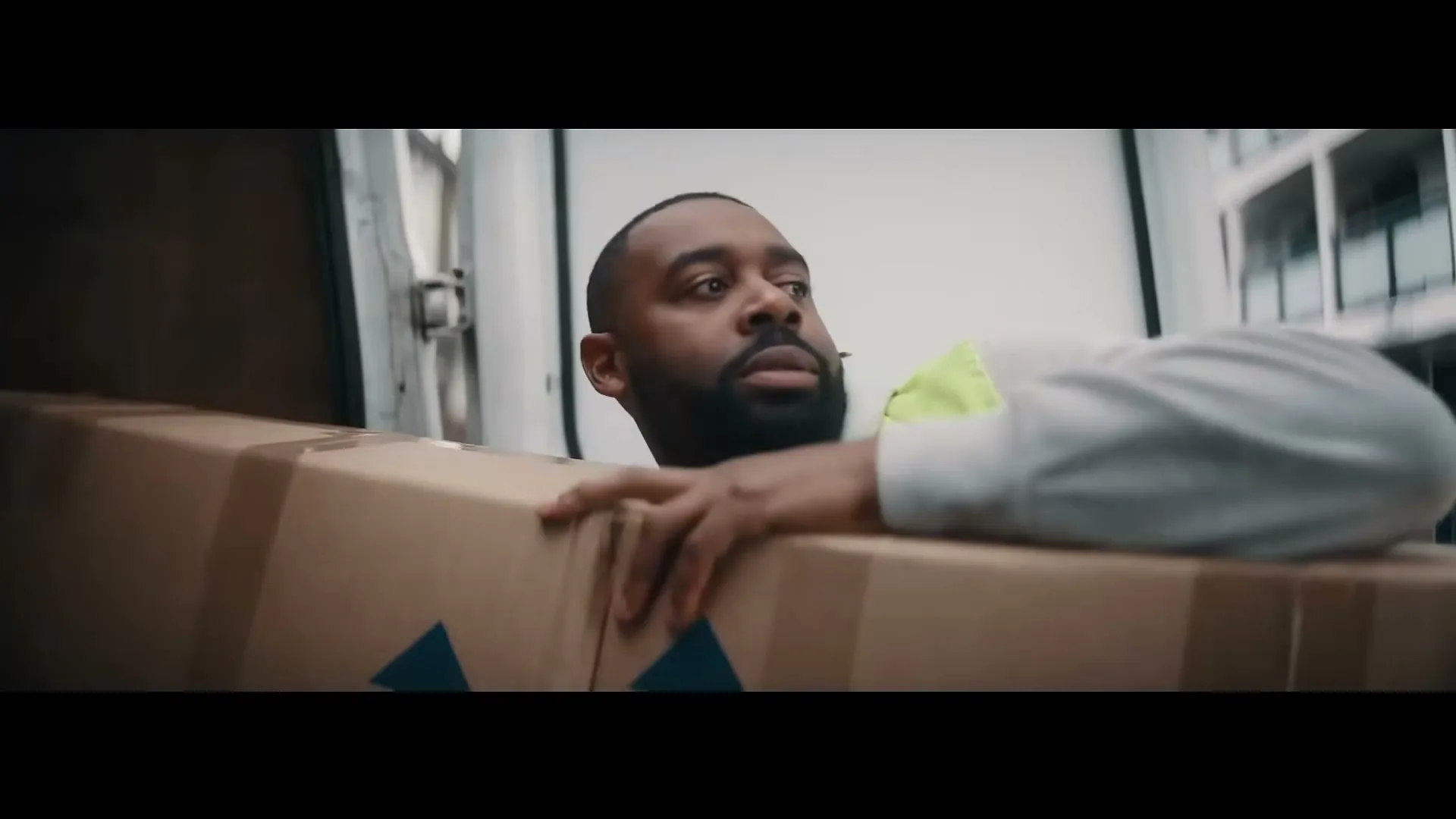 A delivery man with a beard and a high-vis vest is seen from the waist up, looking around nervously from behind a large cardboard box in the back of a van.