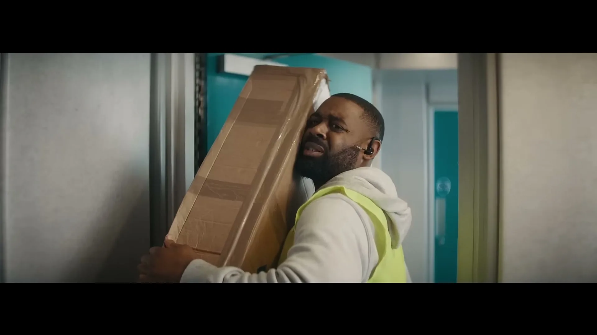 A delivery man in a high-vis vest struggles to carry a large cardboard box through a doorway in an office building, looking strained.
