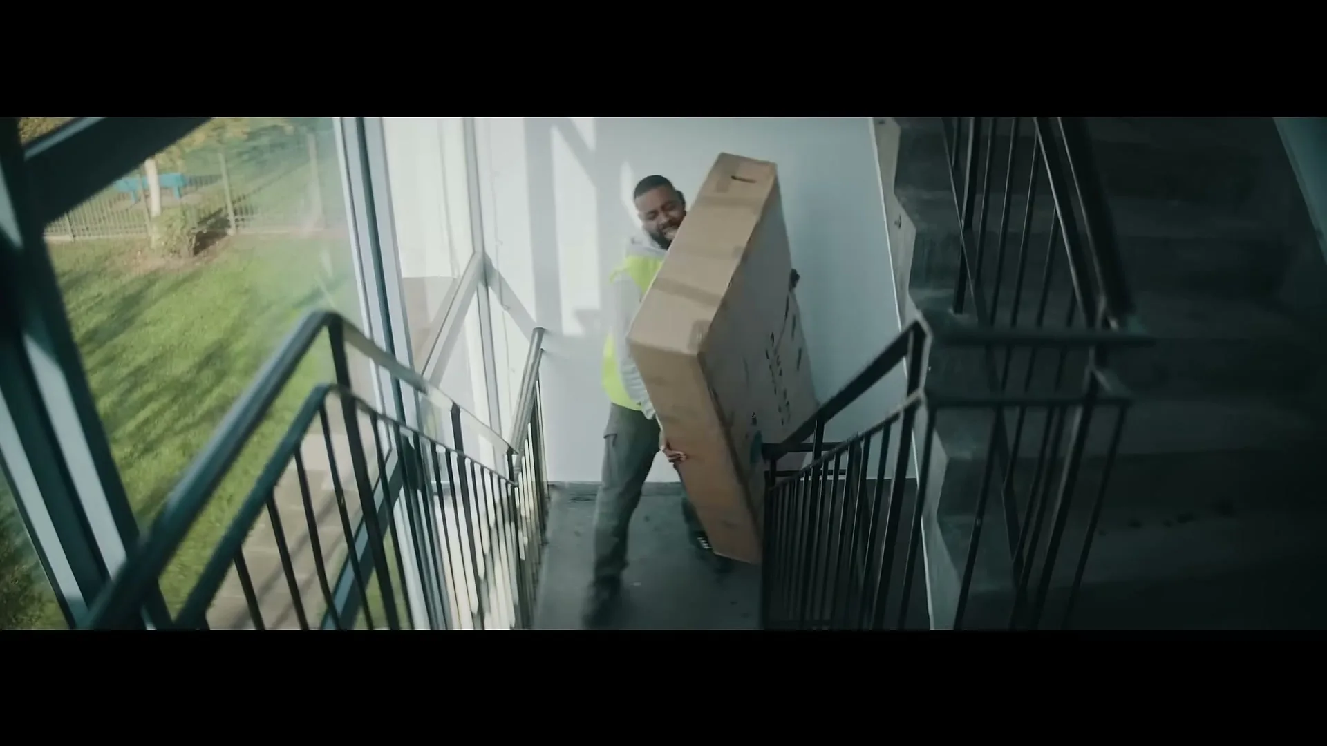 A delivery man in a high-vis vest struggles to carry a large, heavy box up a flight of stairs in an apartment building.