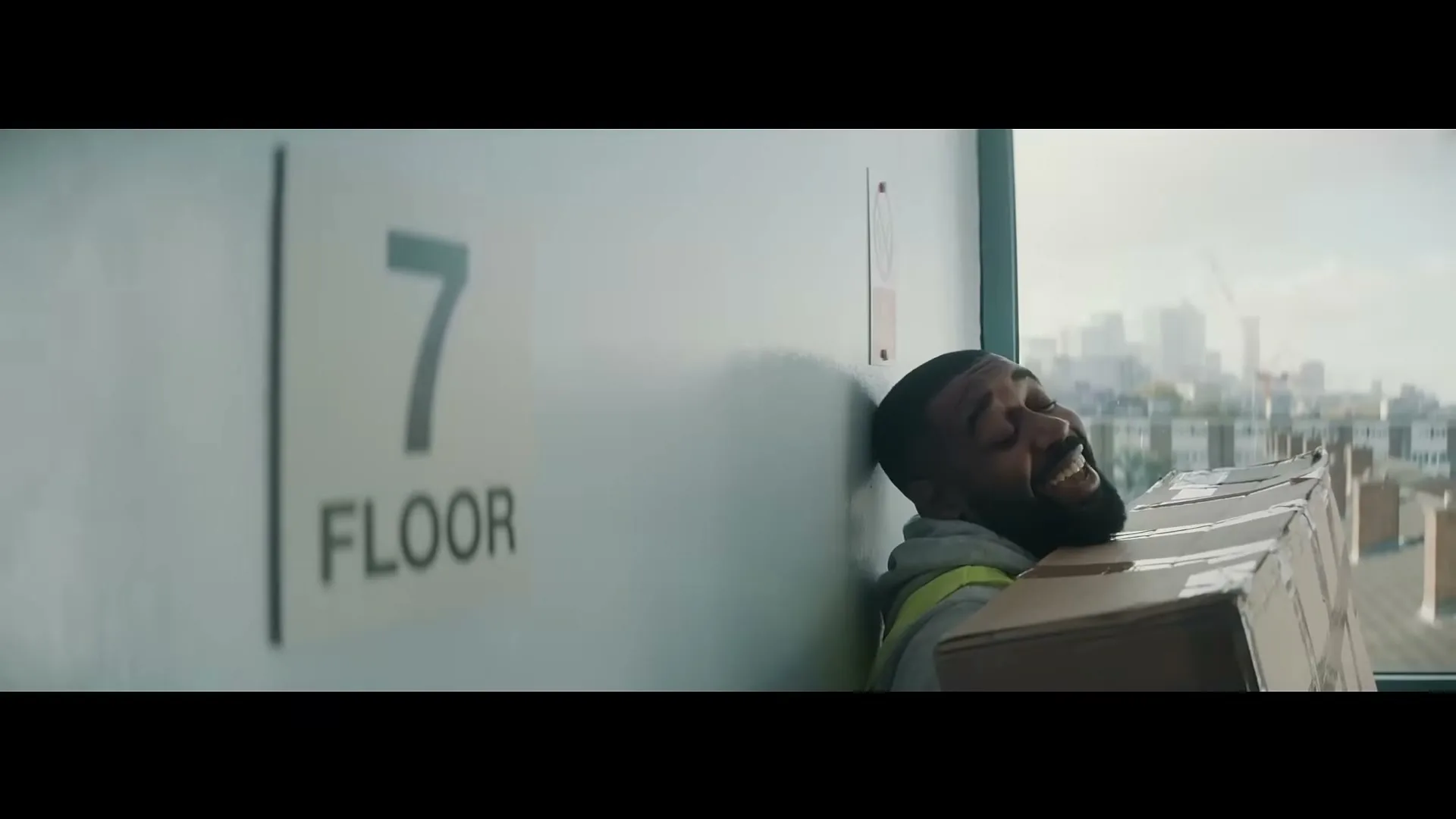 A delivery man in a high-vis vest, exhausted, leans against a wall on the 7th floor, holding a large box, with a city skyline in the background.