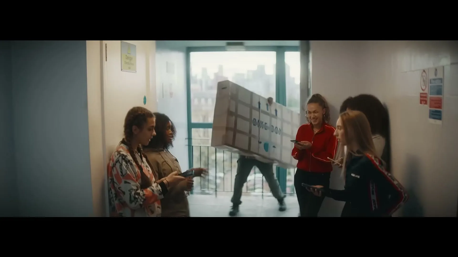 A group of young women stand in a hallway, looking at their phones, as a man struggles to carry a large box through a doorway in an apartment building.