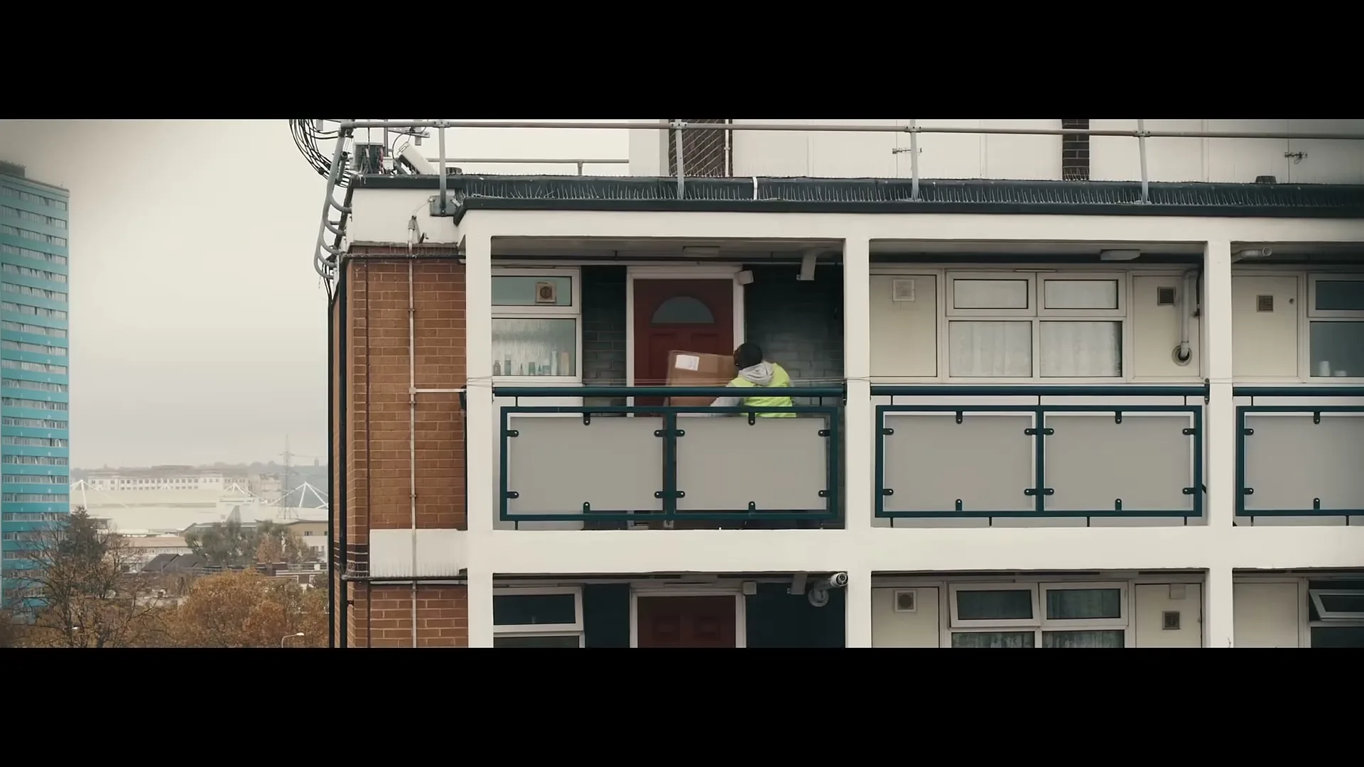 A delivery person in a high-vis jacket attempts to deliver a package to a flat on a balcony of a residential building.