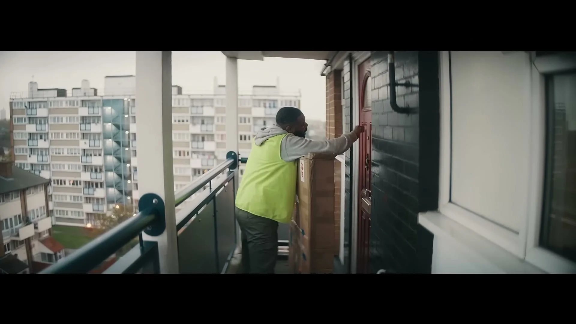A delivery man in a high-vis vest knocks on a red door on a balcony, with apartment buildings in the background.