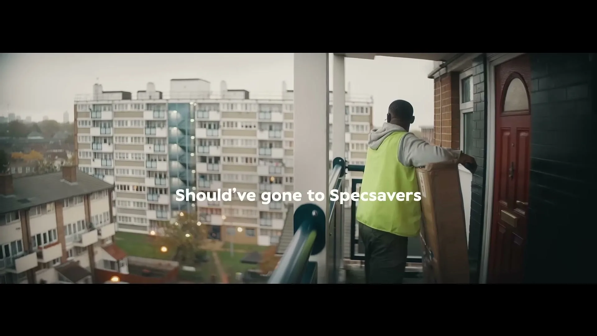 A delivery man in a high-vis vest struggles to fit a large box through a doorway on a balcony overlooking apartment buildings on a cloudy day.