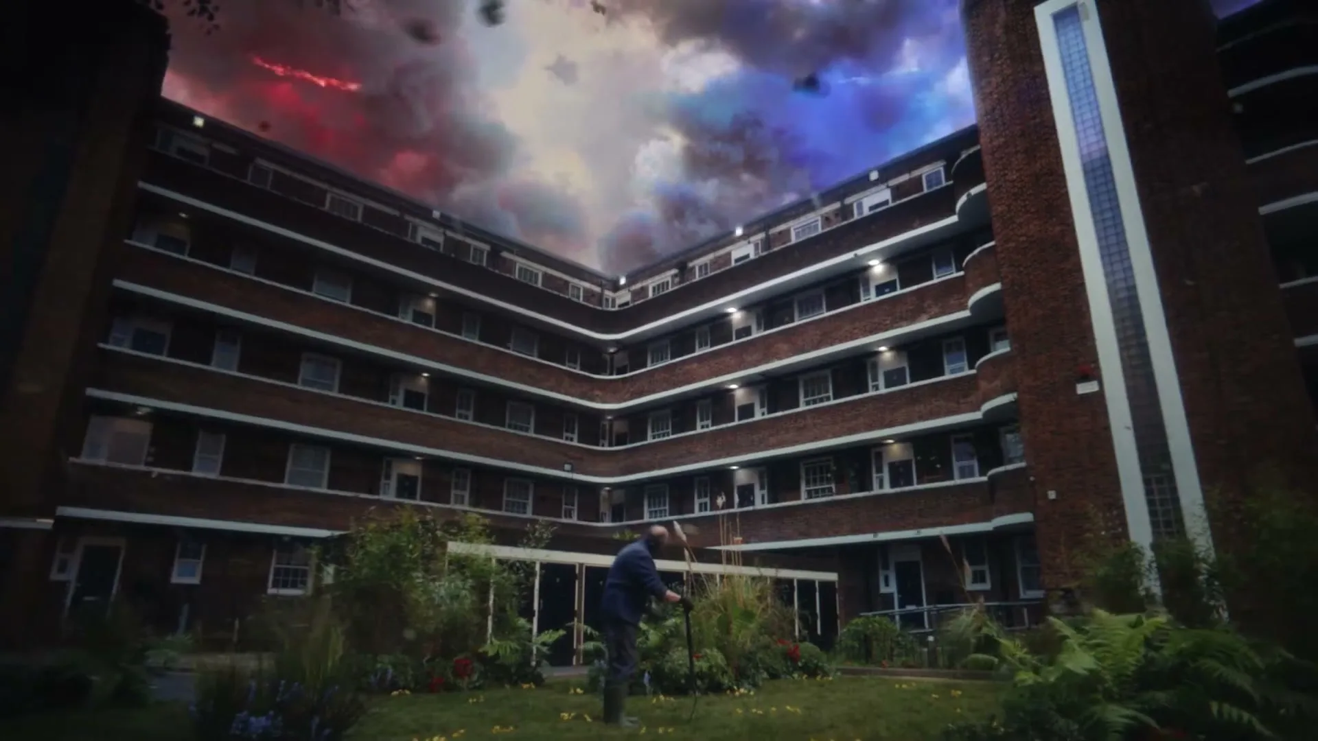 A man in a dark suit tends to a garden in the courtyard of a large brick apartment building, as a dramatic, colorful sky looms overhead.