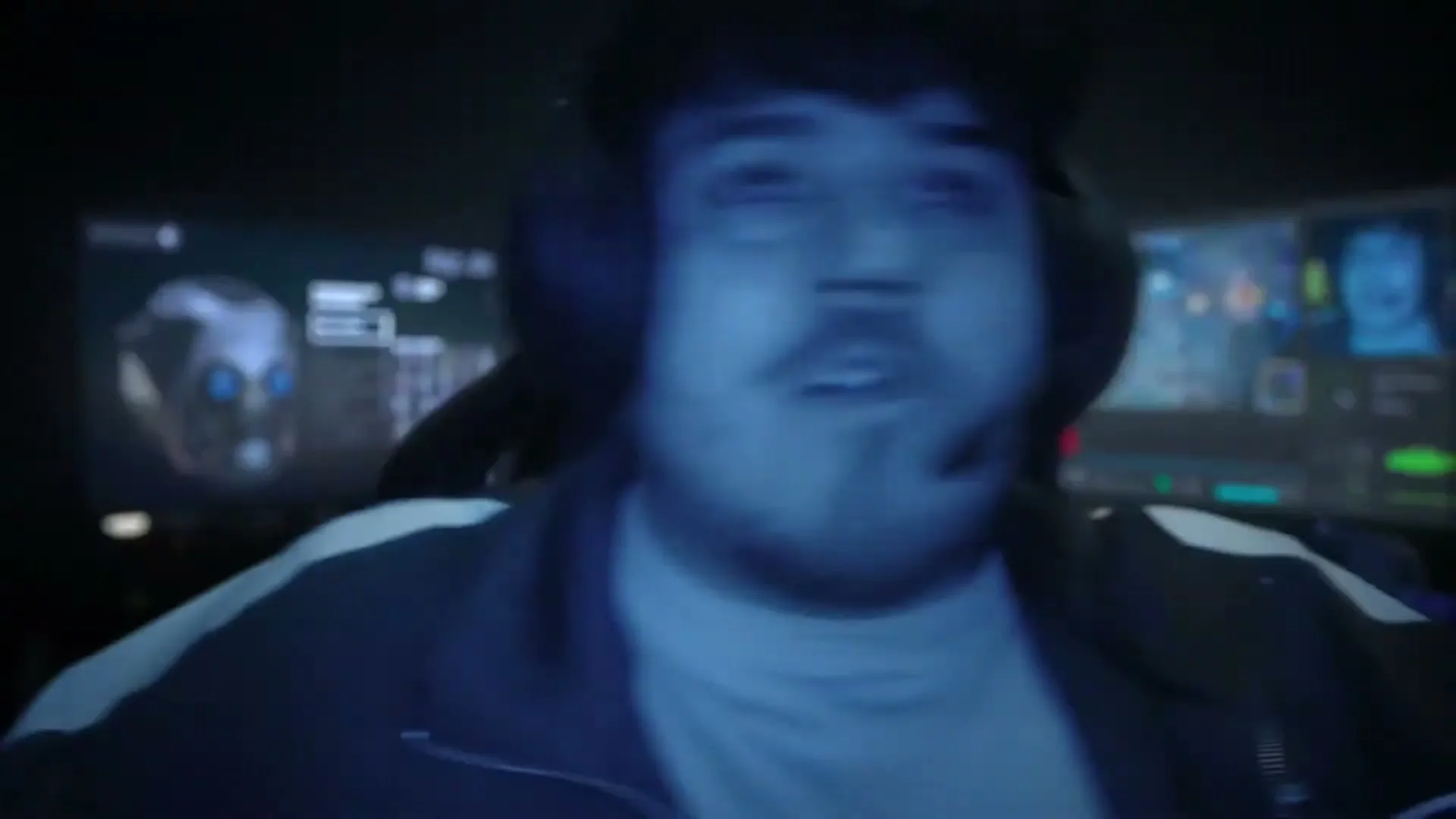 A young man wearing headphones and a tracksuit reacts intensely while playing a video game in a dark room.