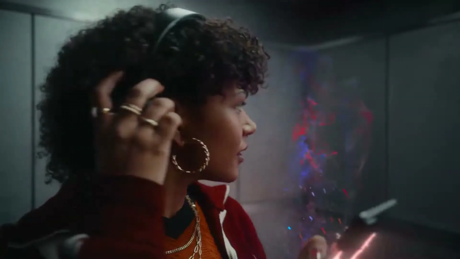 A young woman with curly hair and headphones holds a phone with a glowing red and blue light emanating from it, in an elevator.