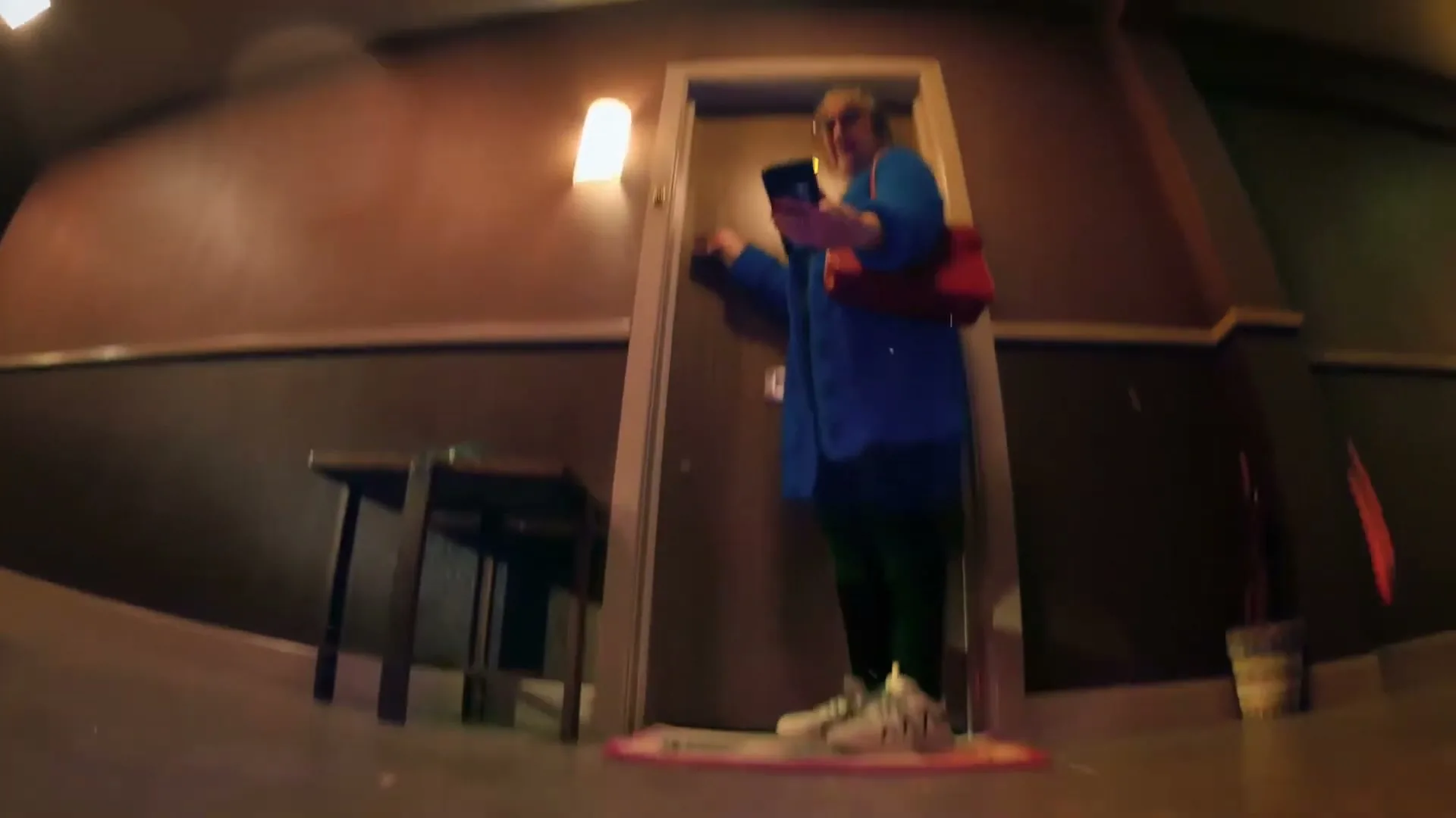 A woman in a blue coat and green pants stands in a hallway, holding a phone and looking around, with a red bag on her shoulder.