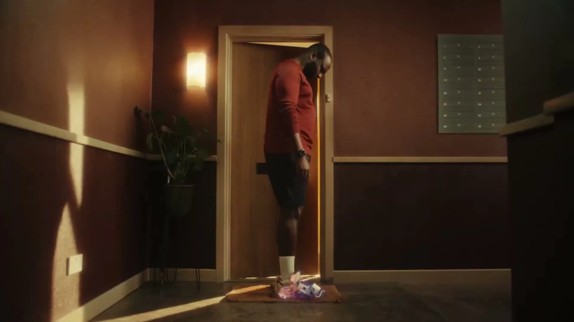 A man in a red shirt and black shorts stands in a doorway, looking down at his glowing white sneakers on a doormat in a dimly lit hallway.