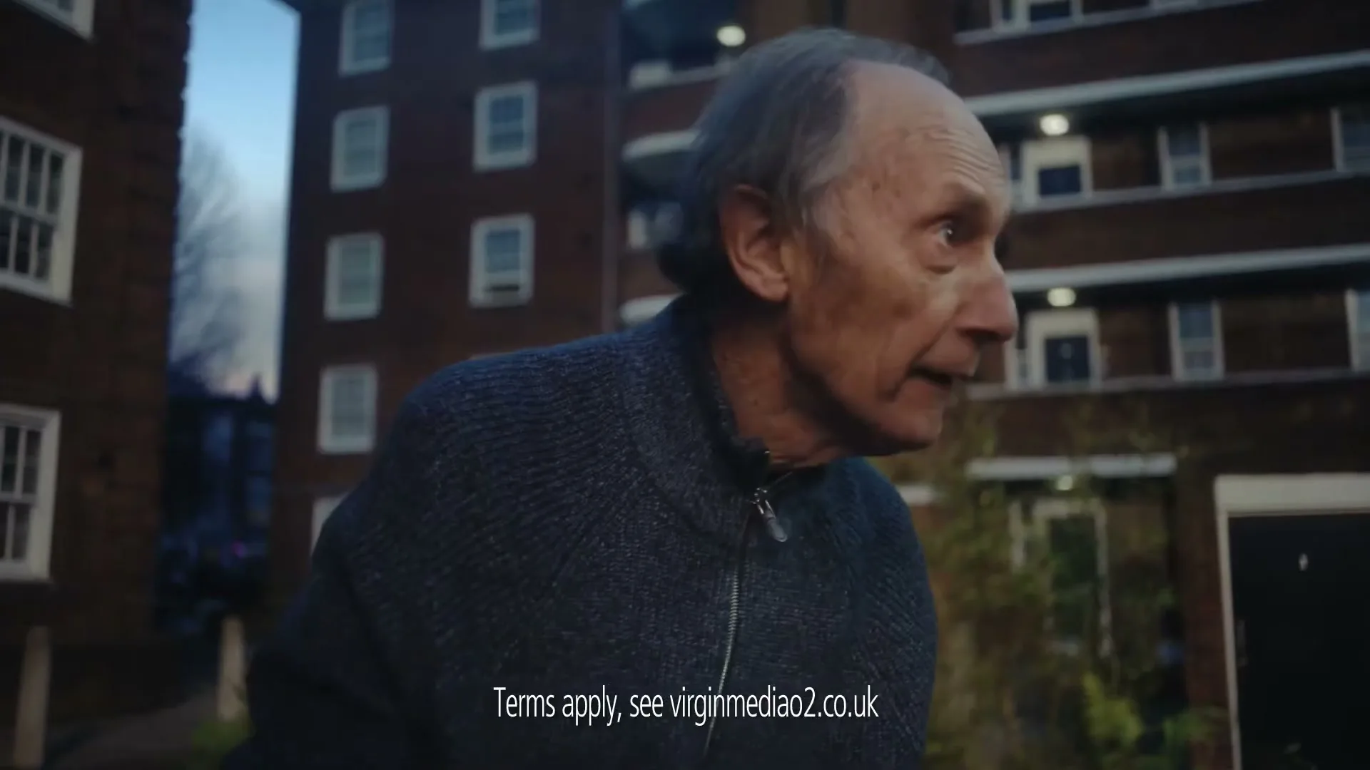 An elderly man in a blue sweater looks around in an urban residential area with brick buildings in the background.