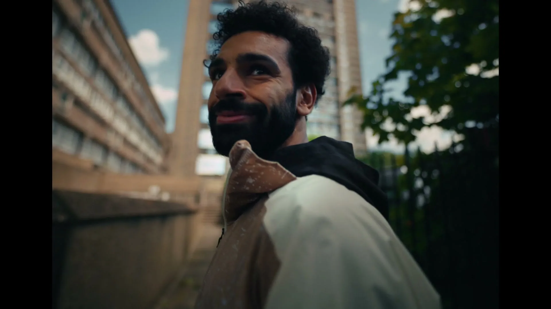 A man with a beard and curly hair, wearing a hoodie, looks back over his shoulder while walking past brutalist buildings and trees.