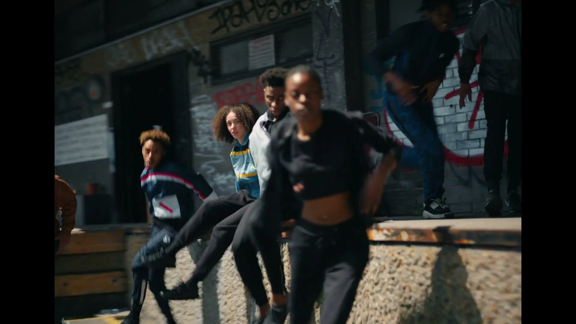 A diverse group of young people in casual streetwear are sitting and then quickly getting up from a concrete ledge in an urban alleyway with graffiti.