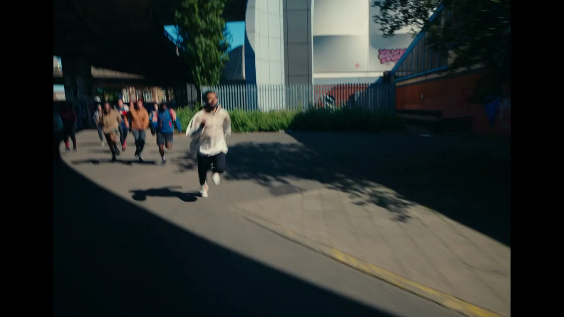 A diverse group of young adults runs down a sun-drenched urban street, with one man in a white hoodie leading the pack.