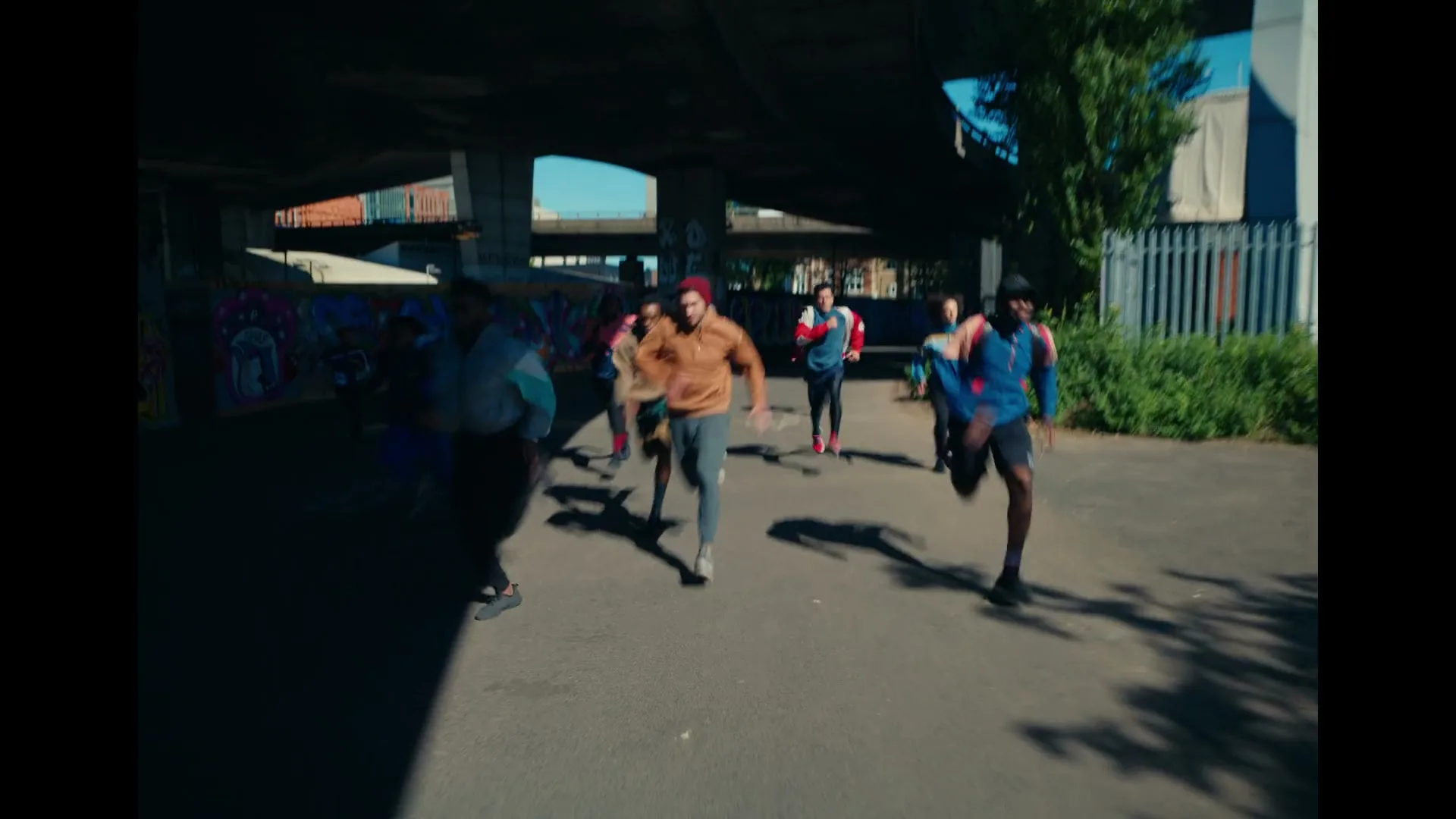 A diverse group of young adults in athletic wear runs together under a bridge on a sunny day, with graffiti visible on the walls.