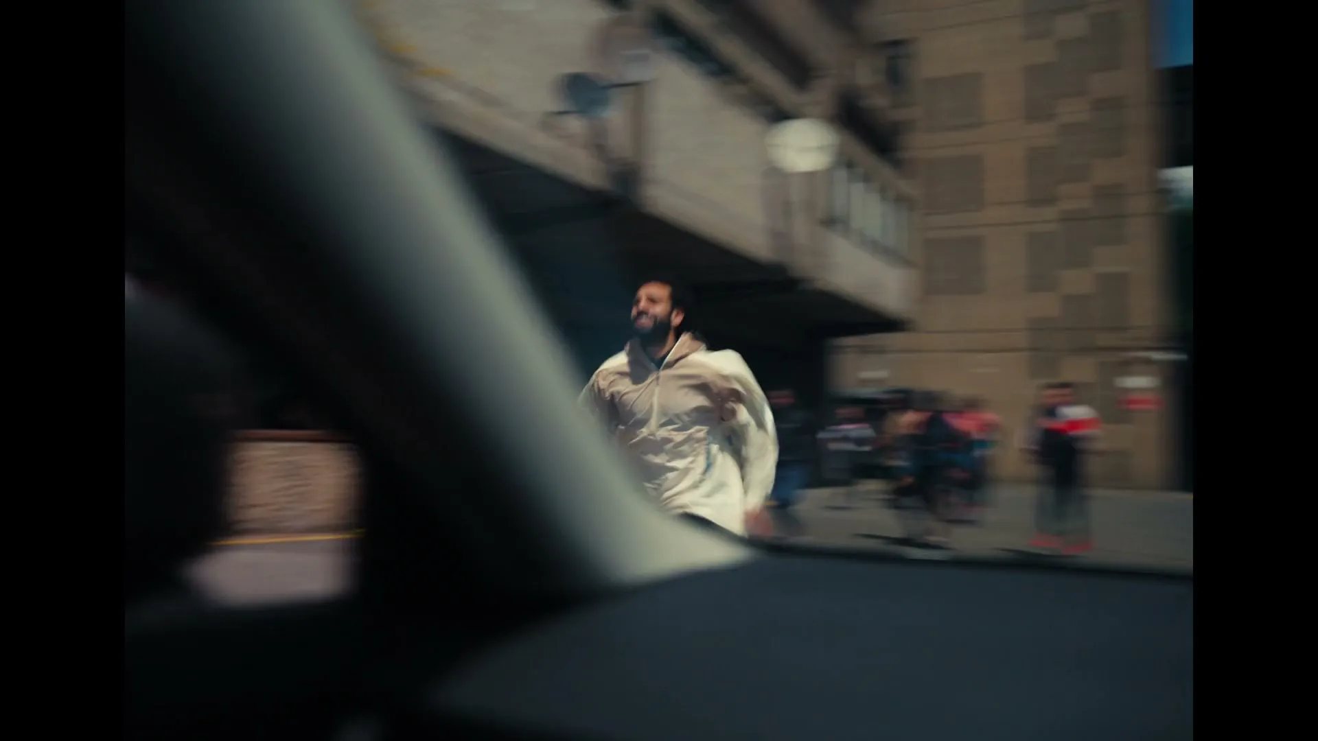 A group of diverse people run towards the camera, which is inside a car, in an urban setting with brutalist architecture.