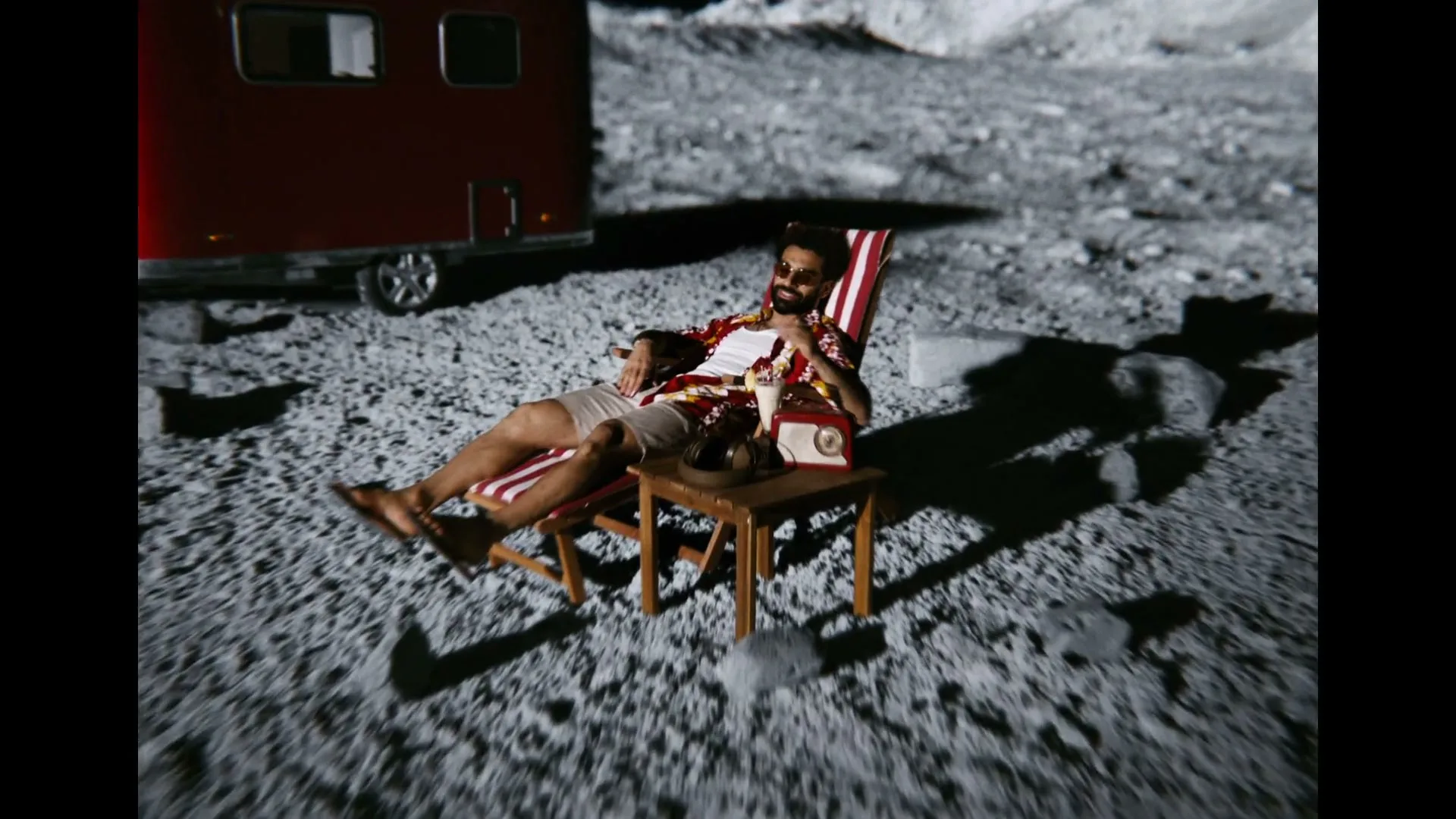 A man with a beard and sunglasses relaxes in a striped deck chair on the moon, holding a drink, with a red camper van in the background.