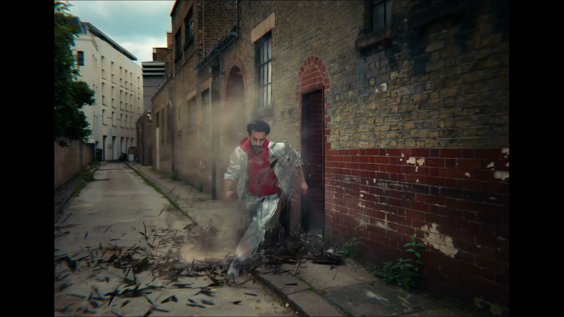 A man in a red hoodie and silver jacket bursts through a brick wall into a narrow alleyway, creating a cloud of dust and debris.