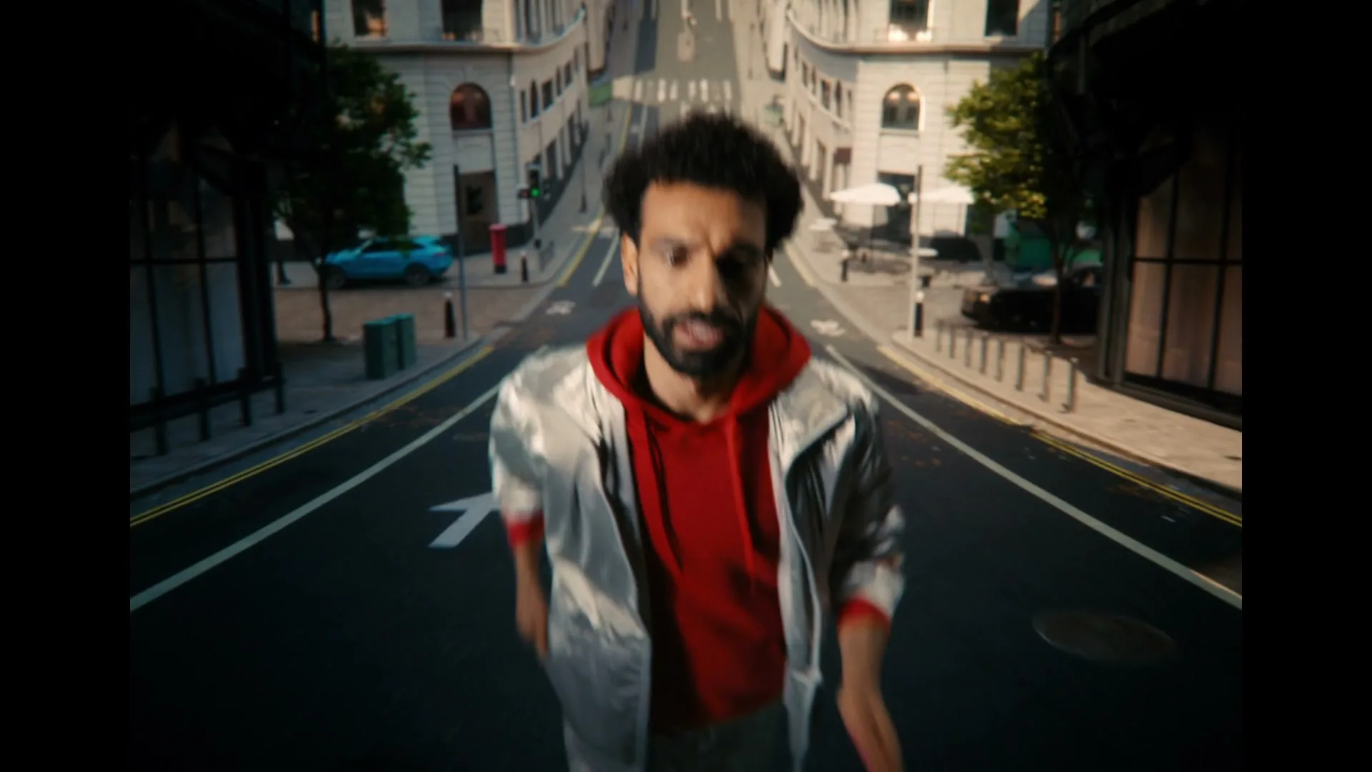 A male athlete with dark curly hair and a beard, wearing a red hoodie and silver jacket, runs towards the camera on a city street, smiling.
