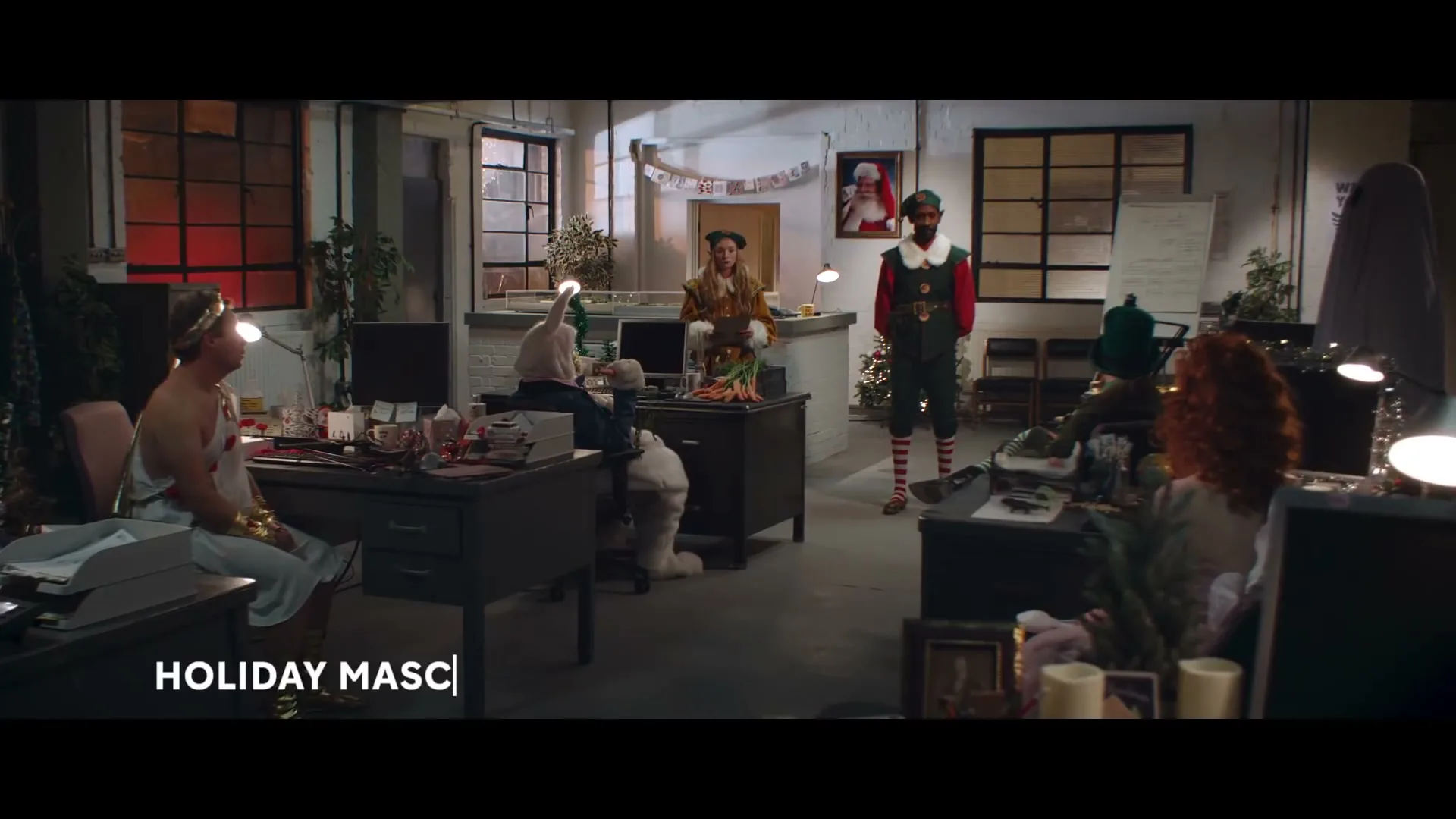 A diverse group of people dressed as holiday mascots are gathered in a dimly lit office, decorated for Christmas, as one person speaks.