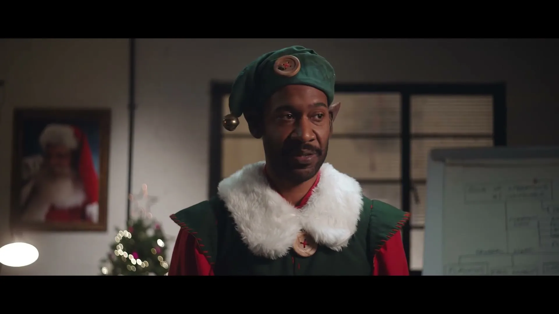 A Black man dressed as an elf with pointy ears and a green hat looks concerned in an office decorated for Christmas.