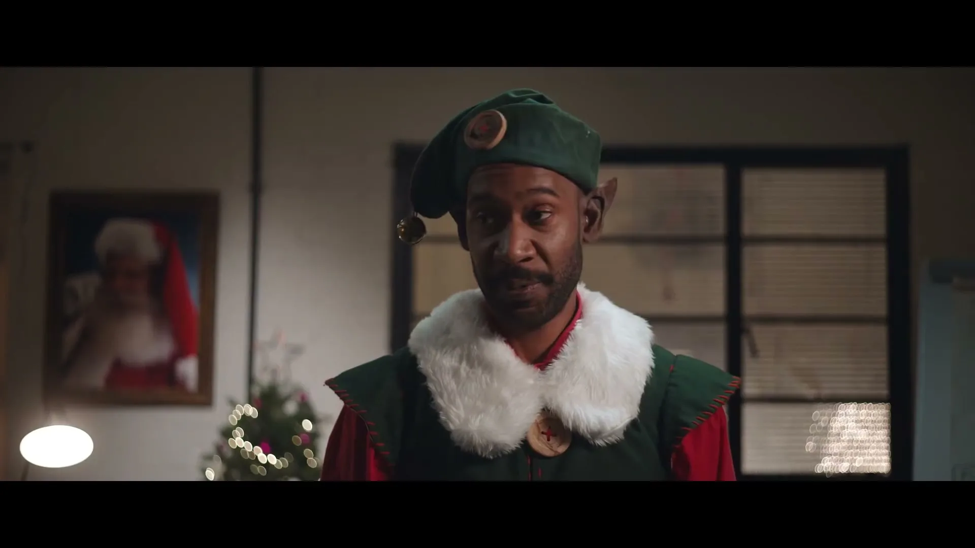 A Black man dressed as a Christmas elf with a green hat and white fur collar looks confused in an office setting with a Christmas tree and Santa portrait.
