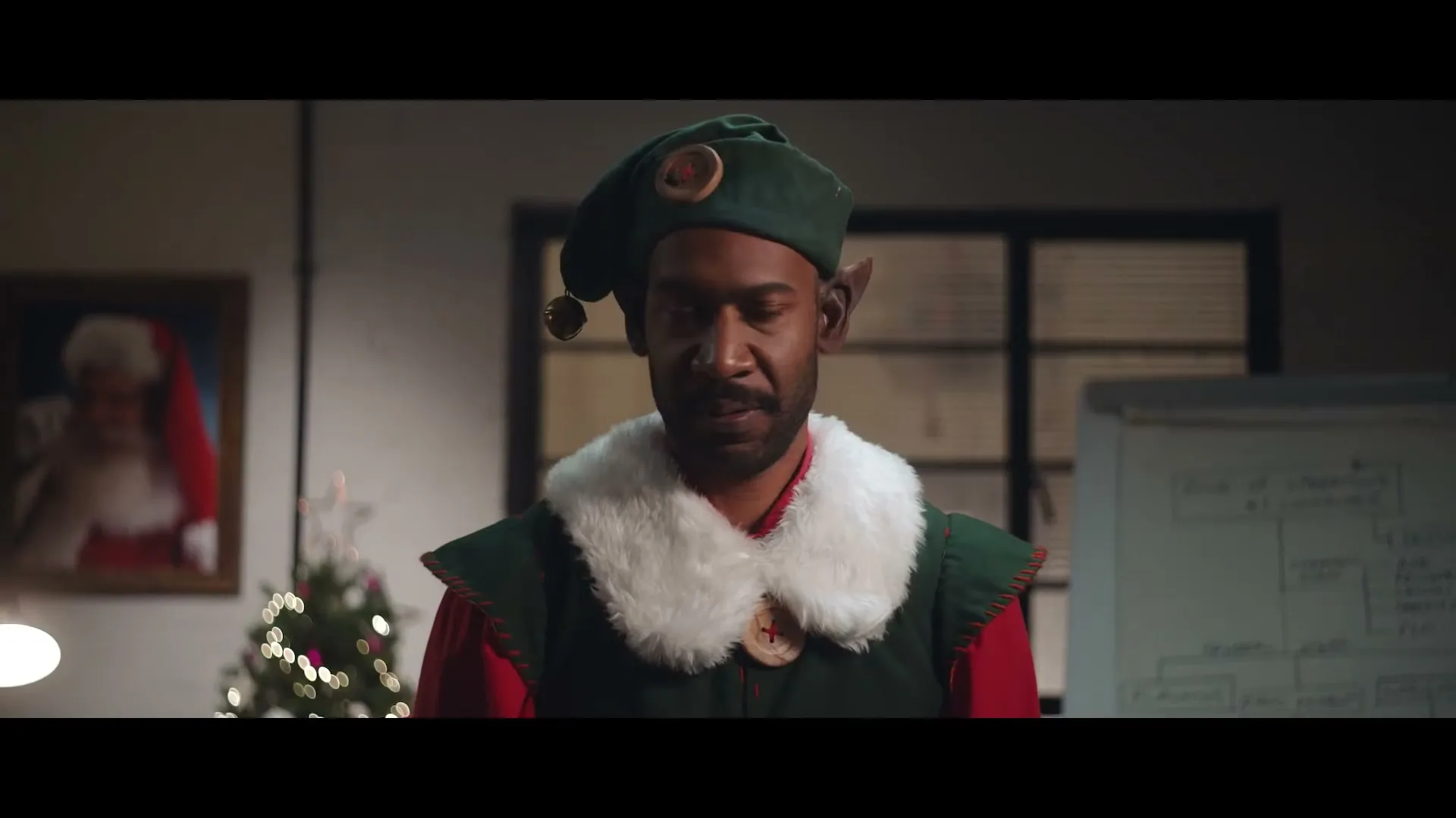 A man dressed as an elf with a green hat and red shirt looks down then up, in an office with a Christmas tree.