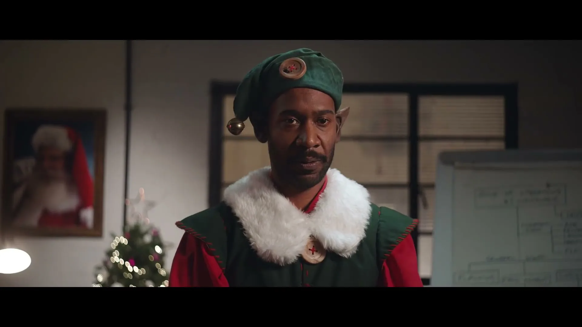 A man dressed as a Christmas elf with pointy ears and a green hat looks directly at the camera in an office setting.