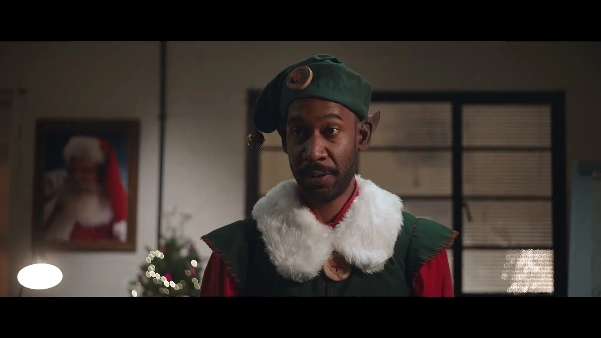 A Black man dressed as an elf with a green hat and white fur collar looks directly at the camera in an office setting.