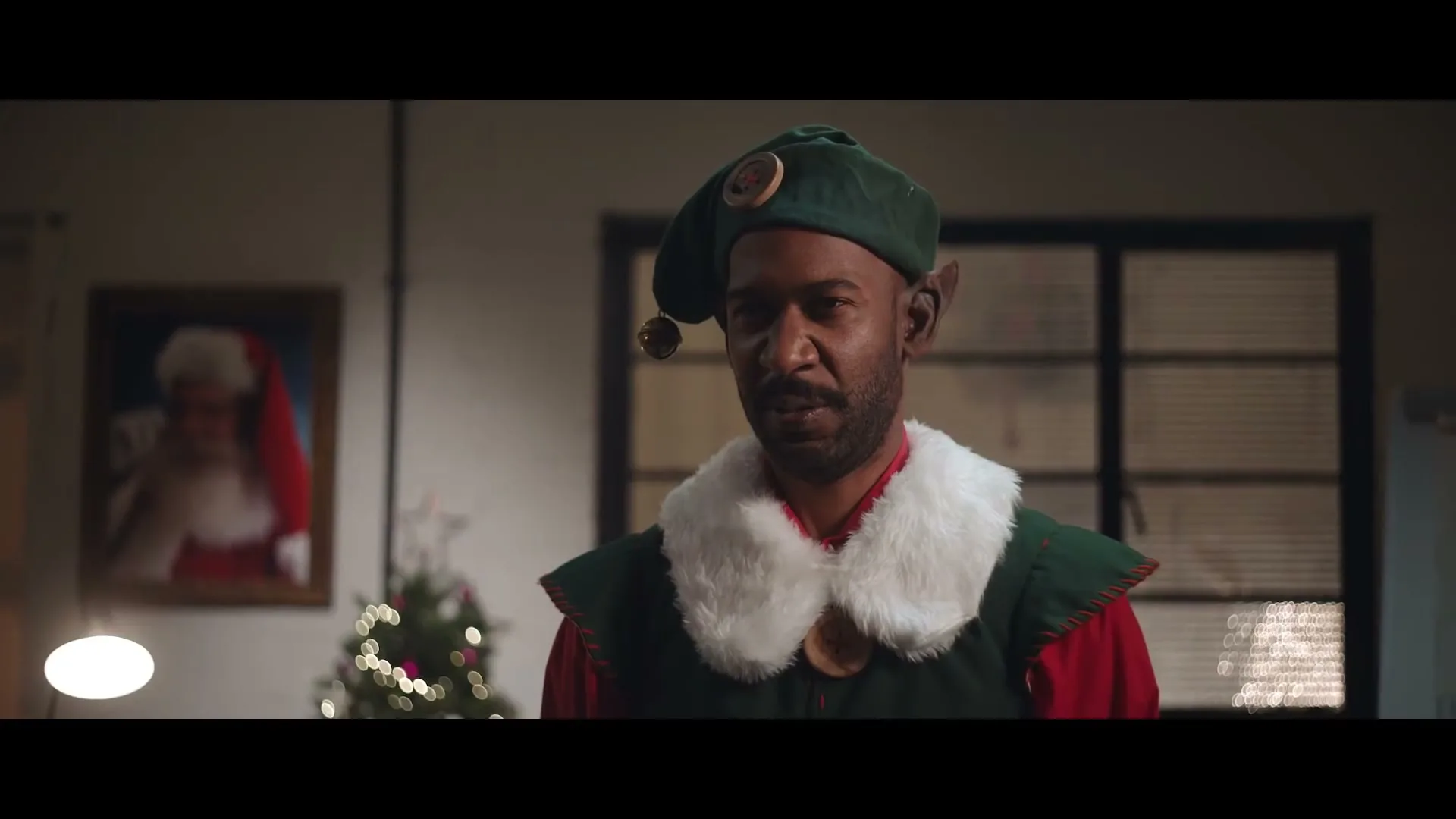 A Black man dressed as an elf with pointed ears looks directly at the camera with a neutral expression in an office decorated for Christmas.