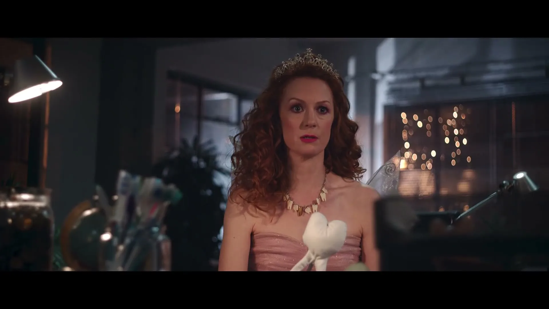 A red-haired woman dressed as a fairy with a tiara and a tooth necklace holds a plush toy, looking concerned in a dimly lit room.
