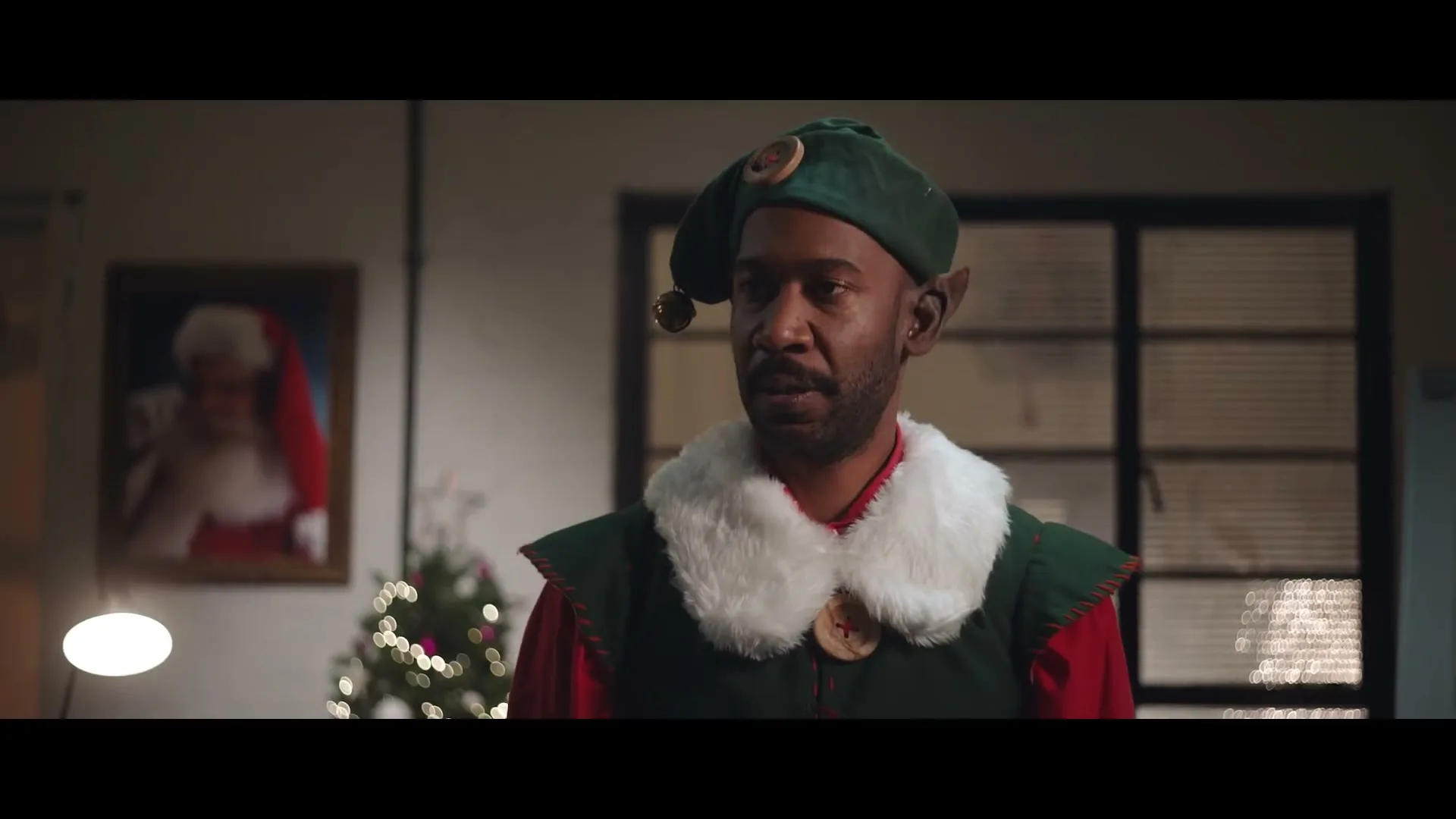 A man dressed as an elf with a green hat and red shirt looks around an office with a Christmas tree and a Santa Claus painting.