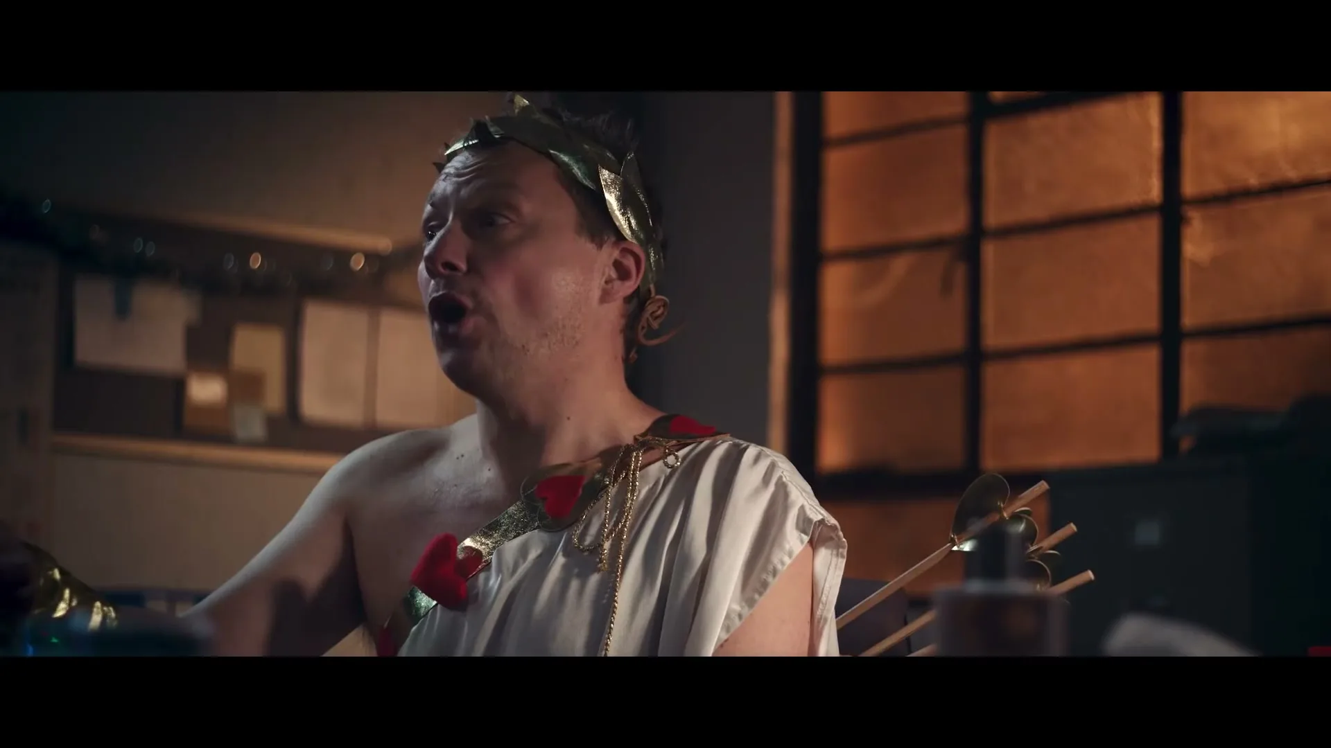 A man dressed as a Roman emperor with a toga and laurel wreath sits at a desk, looking up and speaking in an office.
