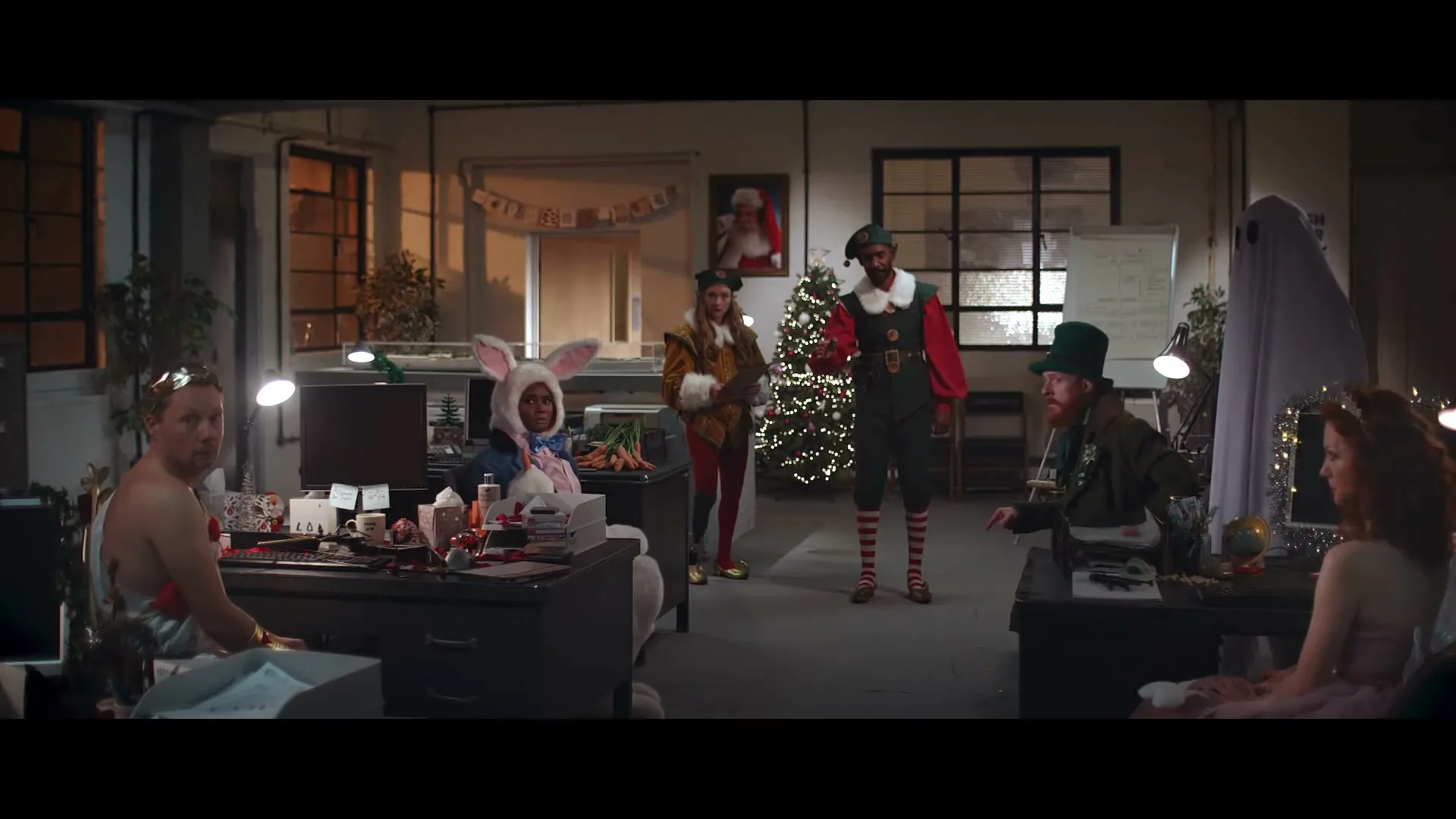 A diverse group of office workers in various festive costumes, including an elf, a ghost, and a Roman emperor, are gathered in a dimly lit office decorated for Christmas.