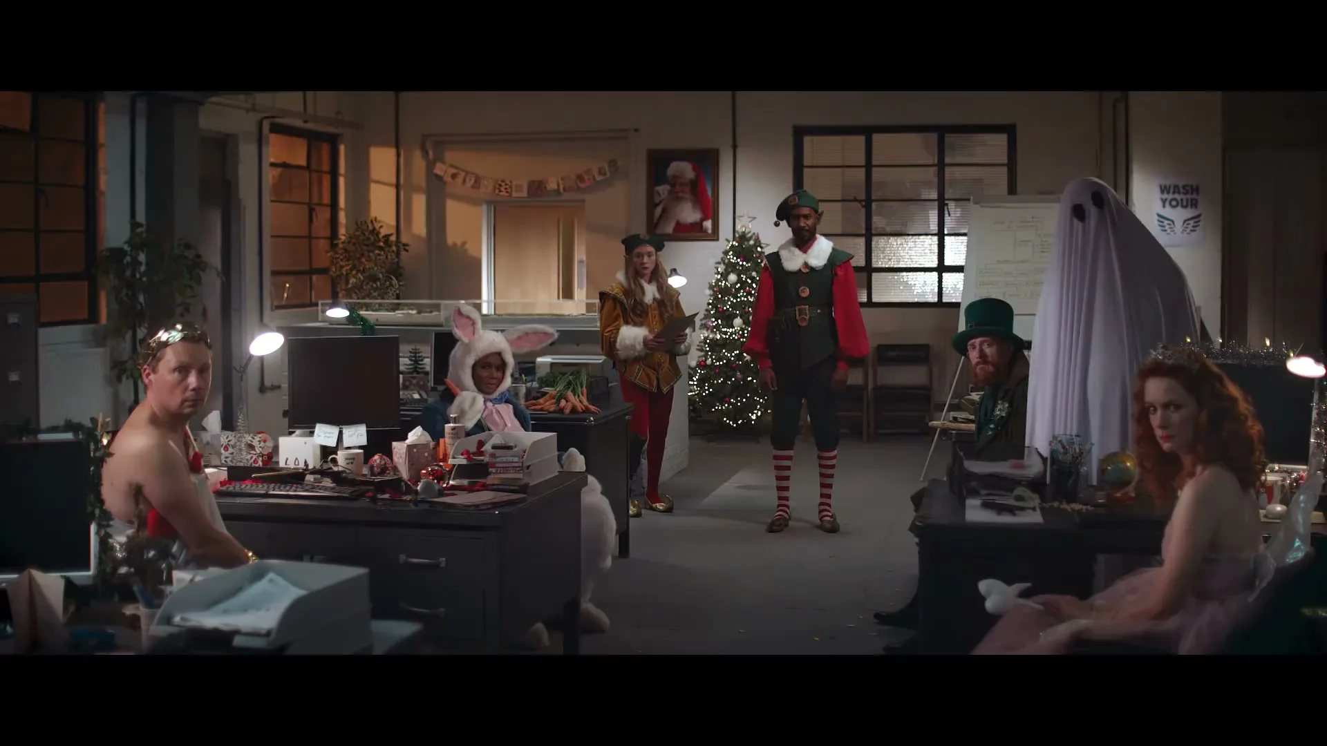 A diverse group of office workers in various Christmas costumes, including an elf, a ghost, and a bunny, are gathered in a festive office setting.
