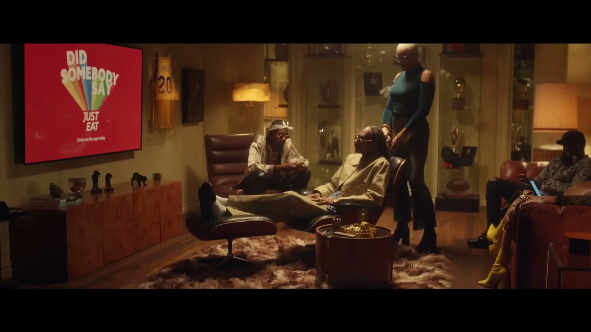 Snoop Dogg, wearing a beige tracksuit, relaxes in a recliner in a dimly lit living room with a group of people, as a woman stands beside him.