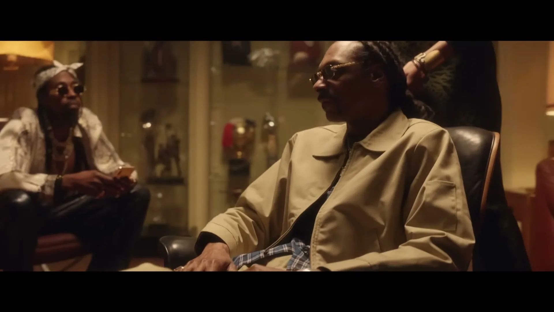 Snoop Dogg, wearing a beige jacket and sunglasses, sits in a leather chair in a dimly lit room, talking to someone off-screen.
