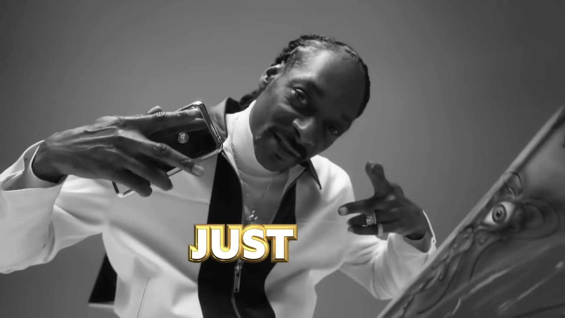 Snoop Dogg, a Black man in his 50s, wearing a white and black jacket, raps and gestures towards the camera in a studio void.