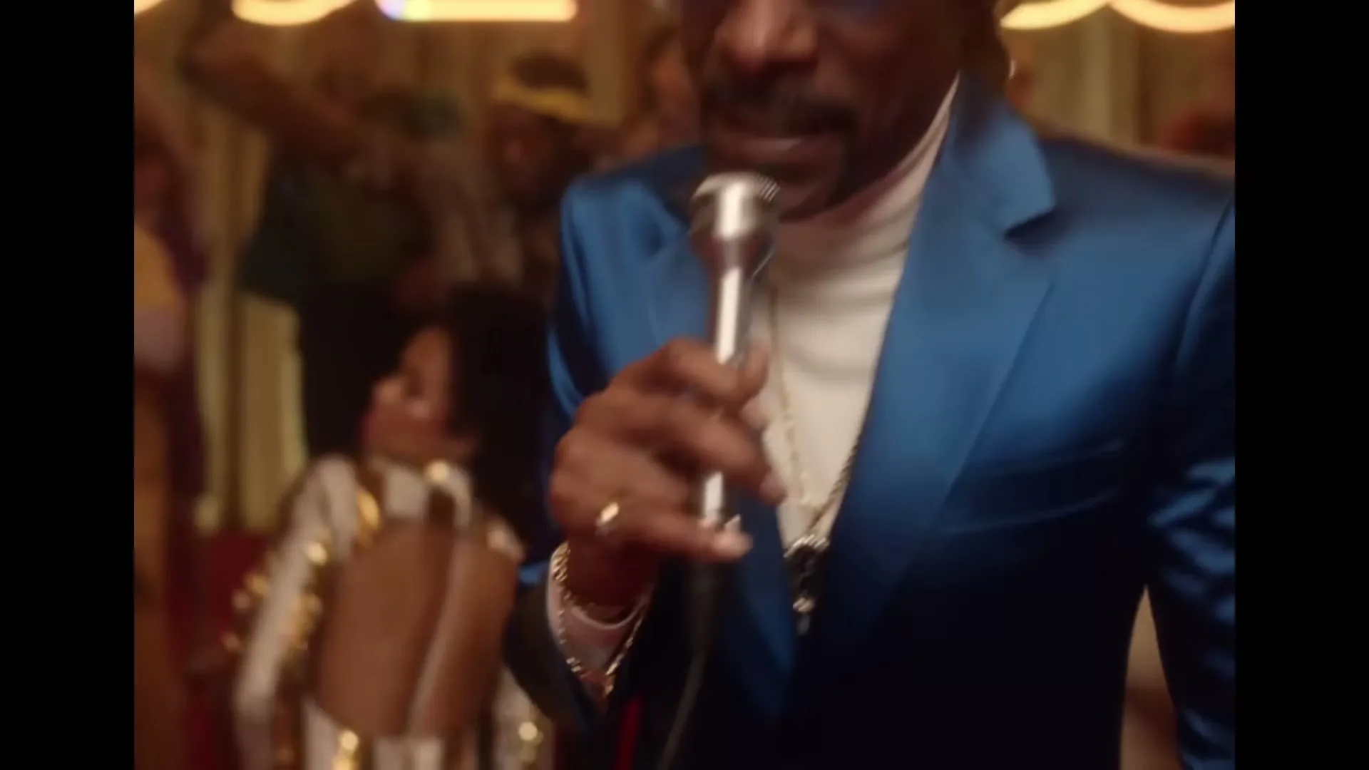 Snoop Dogg, wearing a blue suit and sunglasses, sings into a microphone on a stage with a crowd in the background, with animated text and a dog appearing on screen.