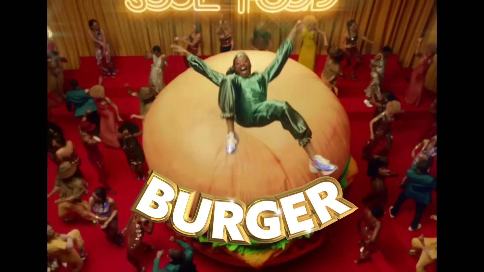 Snoop Dogg, dressed in a green tracksuit, jumps on a giant burger prop while surrounded by dancers on a red carpet stage.