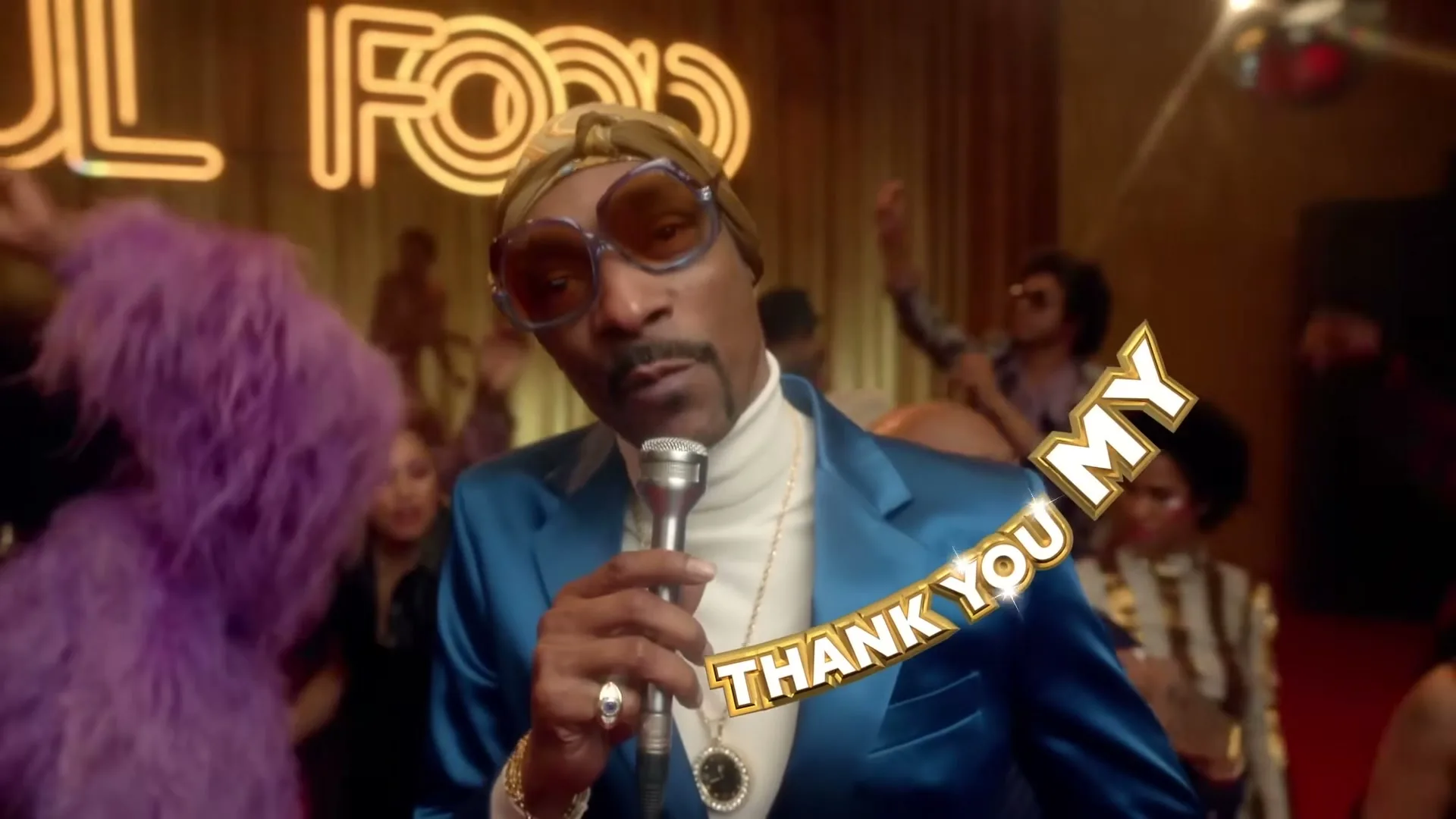 Snoop Dogg, wearing a blue suit, sunglasses, and a headscarf, sings into a microphone on stage at a lively club with a neon "SOUL FOOD" sign in the background.
