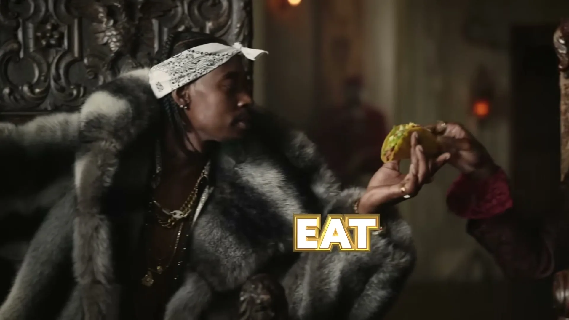 A man in a fur coat and bandana sits on a throne, receiving a taco from another person's hand in a dimly lit, ornate room.
