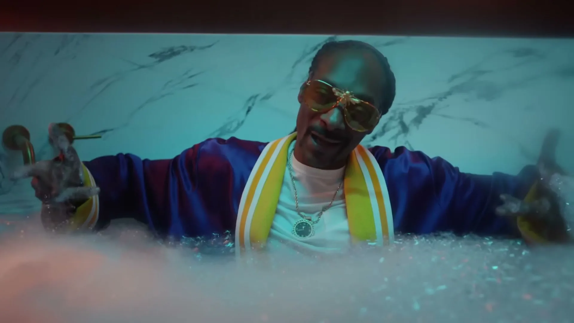 Snoop Dogg, wearing a purple and yellow robe and yellow sunglasses, relaxes in a bubble bath in a luxurious bathroom.