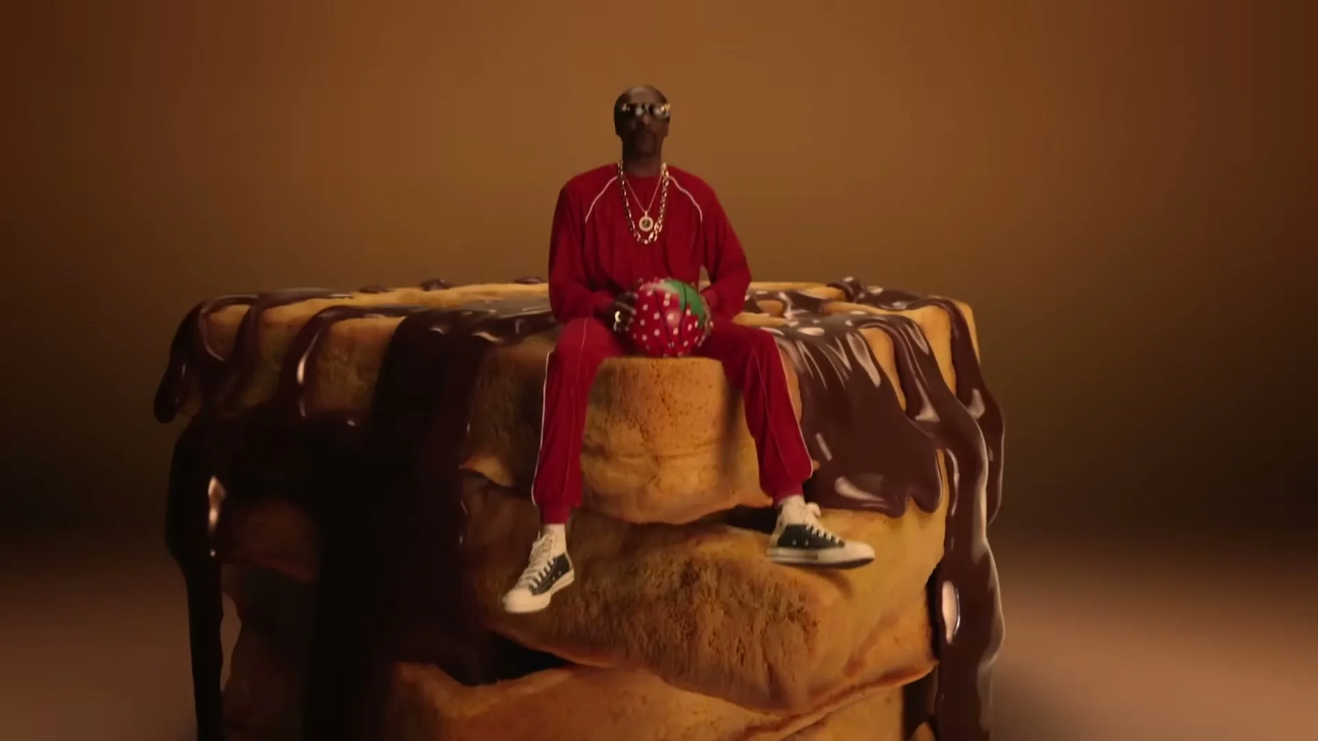 Snoop Dogg, wearing a red tracksuit and sunglasses, sits on a stack of chocolate-covered waffles, holding a strawberry-shaped object in a studio.